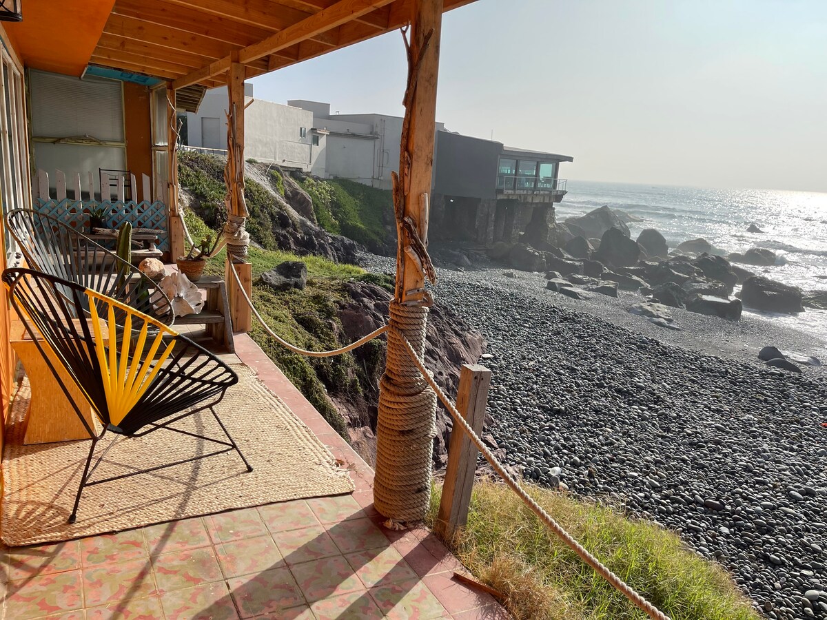 A cozy outdoor seating area features a vibrant black and yellow chair set on a tiled patio. The view includes tranquil ocean waves lapping against pebble rocks. Sunlight casts a warm glow, enhancing the connection between the dwelling and the natural surroundings.