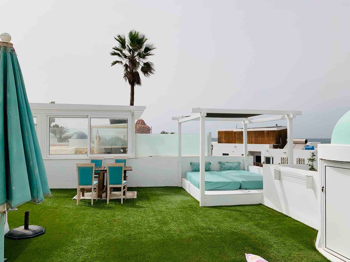 A spacious rooftop terrace is surrounded by white walls, featuring a large lounge bed with soft cushions and a dining set with turquoise chairs. A vibrant umbrella offers shade, while grass-like flooring adds a natural touch. Palm trees are visible in the background against a cloudy sky.