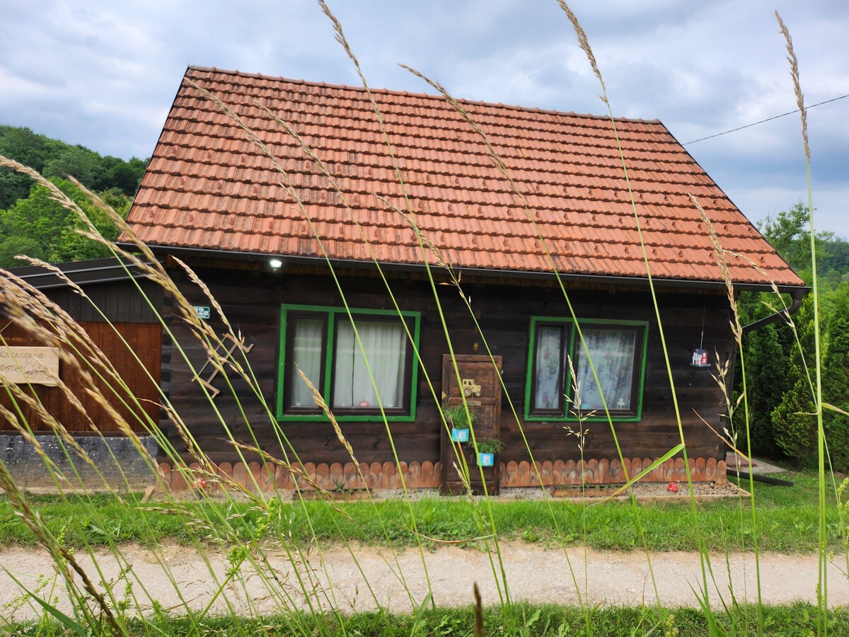 The charming wooden cabin, featuring a traditional sloped roof with terracotta tiles, is surrounded by lush greenery. Large windows provide glimpses of the cozy interior, while a stone pathway leads up to the entrance. The rustic architecture is complemented by natural grass softly swaying in the foreground.