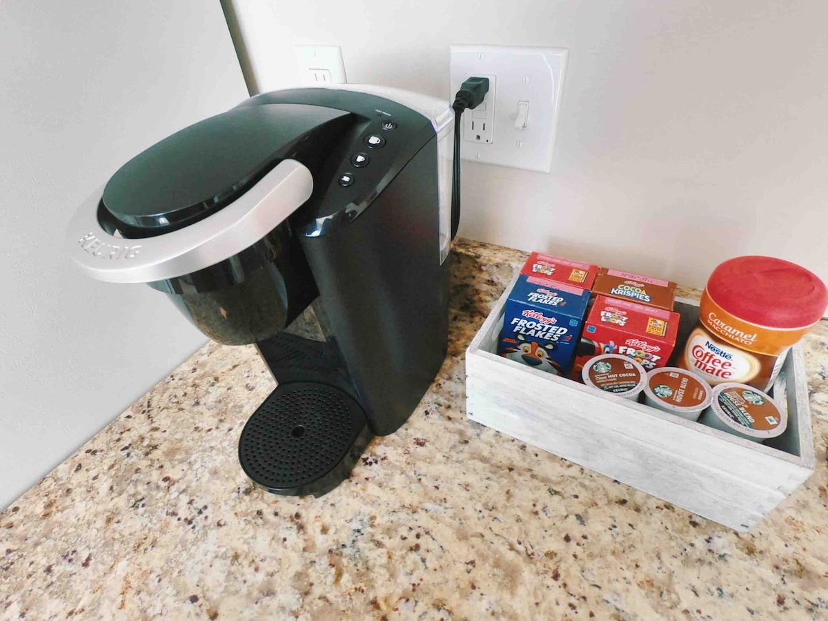 A modern coffee maker is positioned on a speckled countertop, with a selection of coffee and cocoa pods neatly arranged in a wooden tray beside it. The tray includes various flavors and a jar of spread, offering a convenient beverage option.