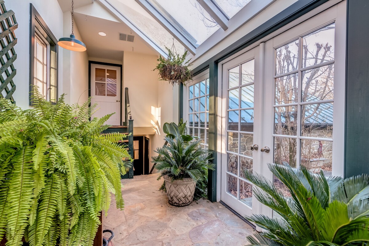 A sunlit sunroom features an array of green plants in decorative pots, set against a backdrop of large glass doors leading outside. The stone flooring complements the serene environment, while natural light filters through the skylights above.