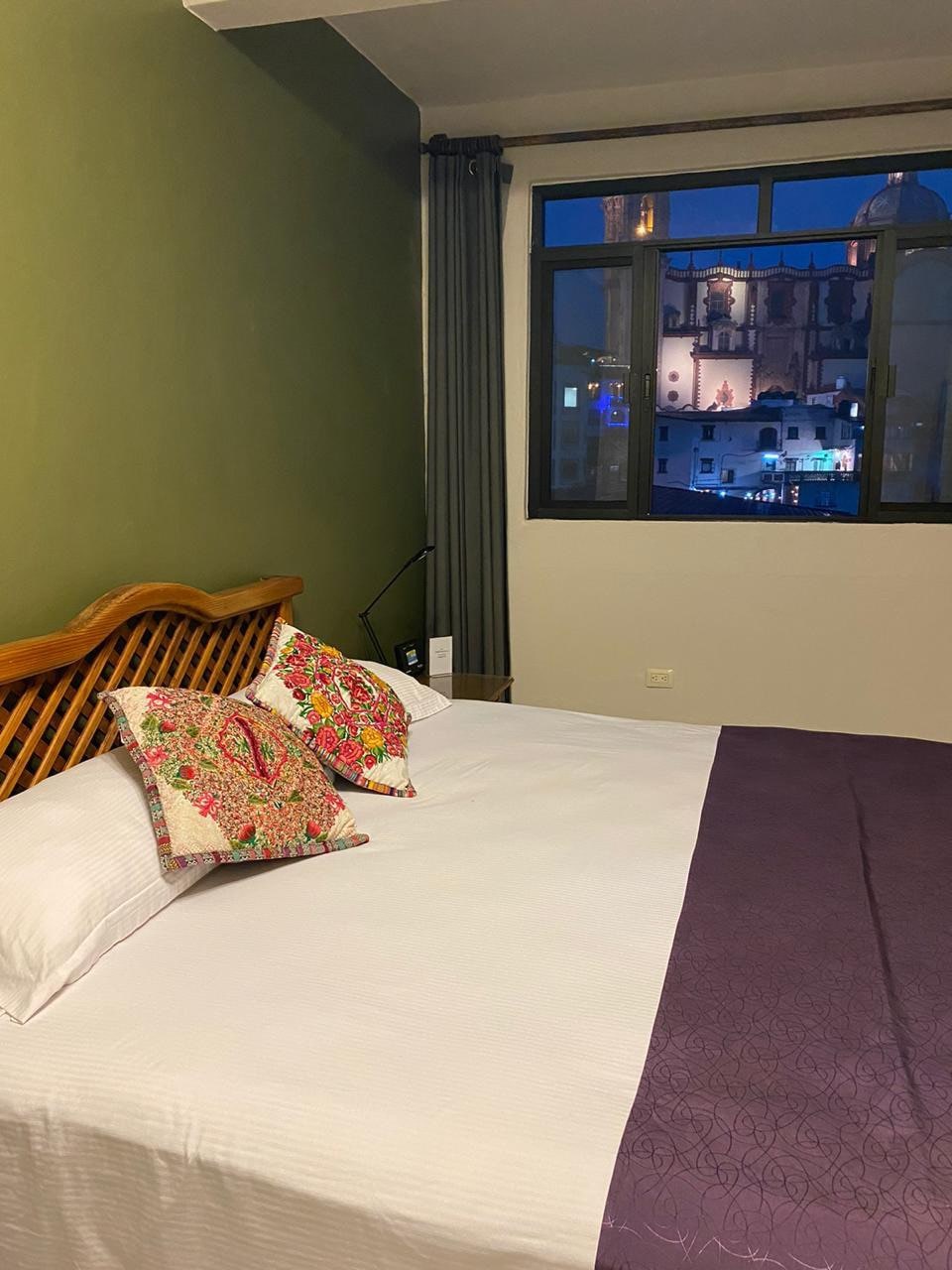 A spacious bed is placed against a green wall, adorned with decorative cushions featuring colorful floral patterns. Light filters through a large window, showcasing views of a cityscape illuminated at night, with architectural details visible from the nearby Cathedral.