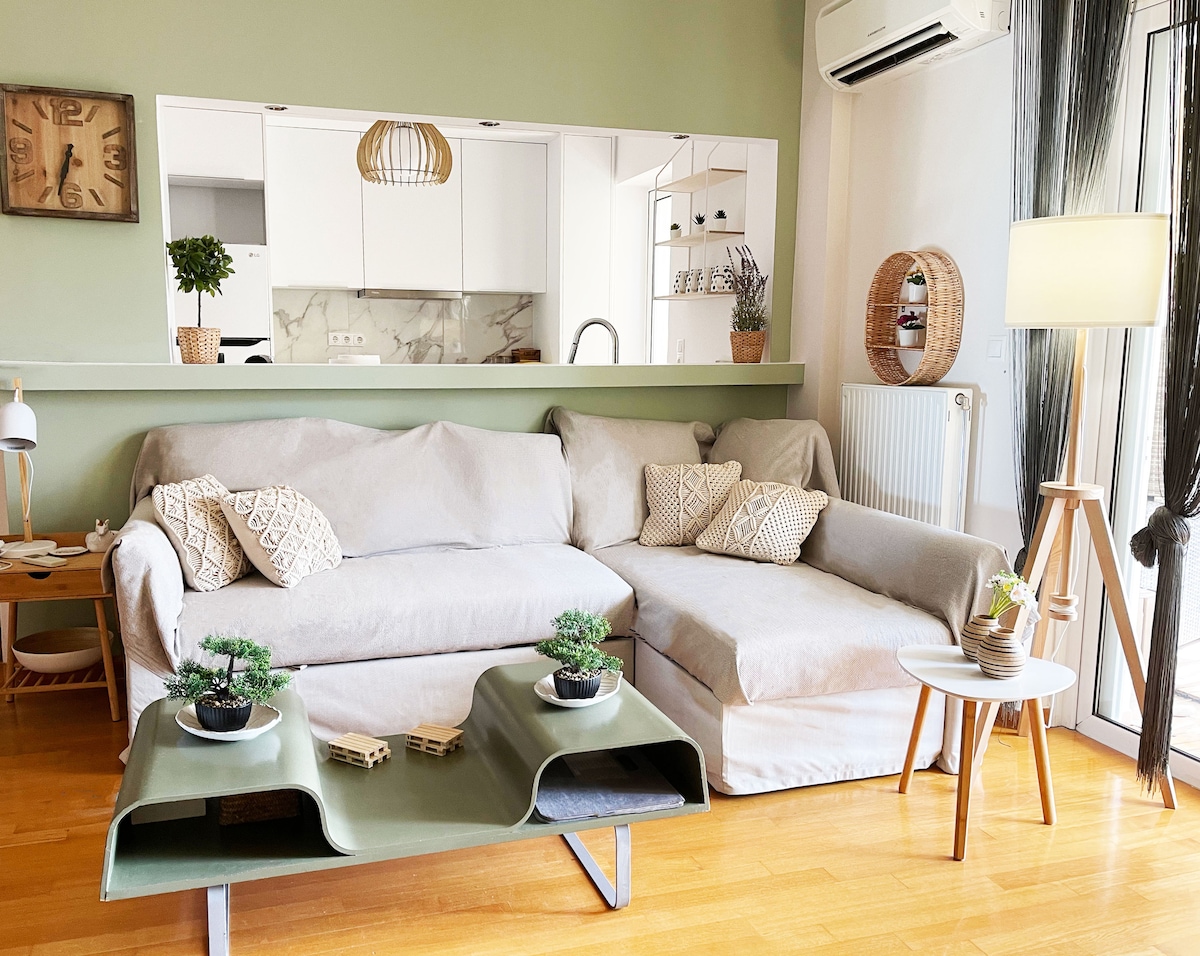 A modern living area features a spacious sectional sofa with soft cushions and decorative throw pillows. A sleek, green coffee table holds small plants, while natural light floods the room from nearby windows. A mirror reflects the open kitchen area, enhancing the interior's bright ambiance.