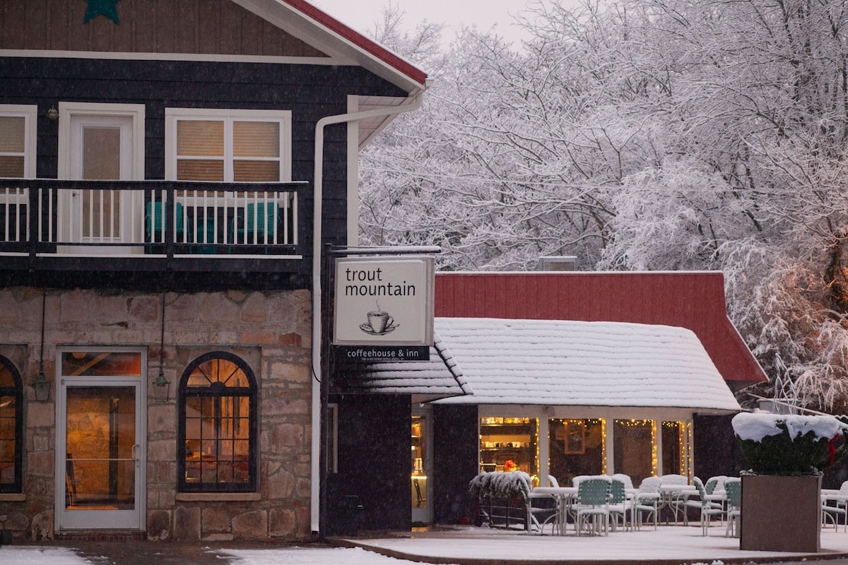 A charming building features a stone and wood exterior, with large windows and a welcoming balcony. Snow blankets the scene, adding a serene touch. The sign for Trout Mountain Coffeehouse & Inn is prominently displayed, while outdoor seating is arranged on a covered patio.