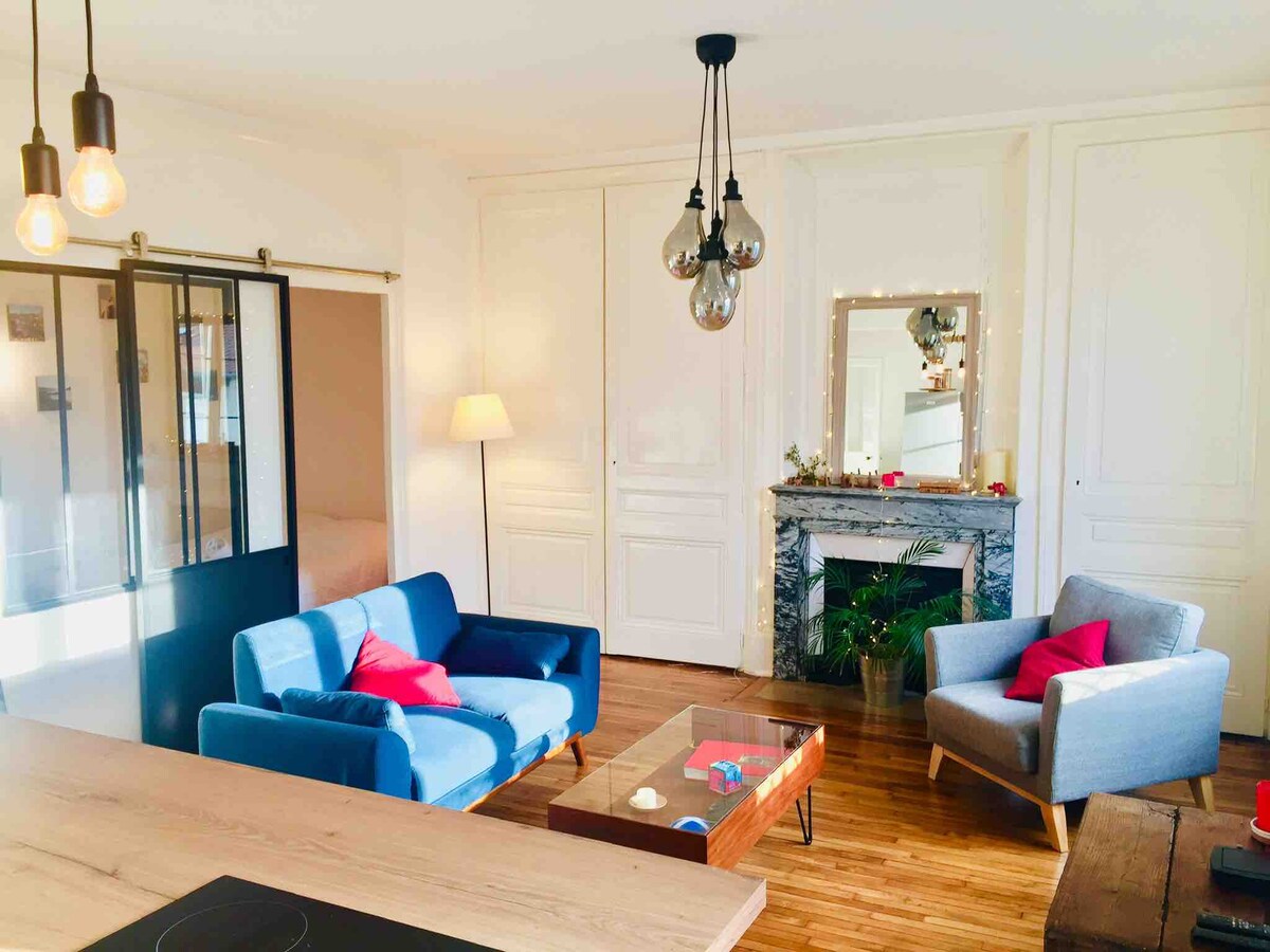 A living area is presented with a blue sofa and a gray armchair, featuring red cushions for added comfort. A wooden coffee table sits between them, and a marble fireplace is visible, adorned with decorative elements. The space is illuminated by pendant lights, and natural light enters through large windows.