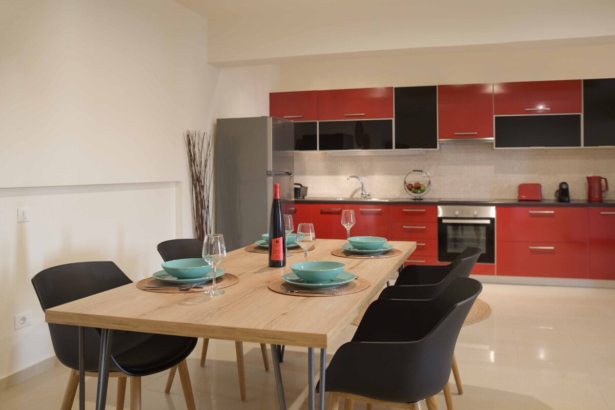 A modern kitchen is captured, featuring red cabinetry and stainless steel appliances. A wooden dining table is set with turquoise bowls and glassware, inviting shared meals. A sleek refrigerator stands alongside the kitchen, enhancing the functional use of space.