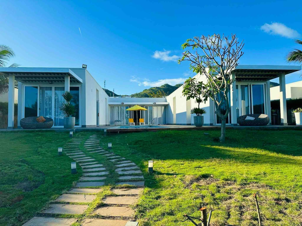 The villa's exterior features a modern design with two symmetrical buildings flanking a central area. A pathway of stepping stones leads through a lush green lawn, while large glass doors reveal a sunlit terrace with outdoor seating. Palms and decorative plants enhance the inviting landscape.