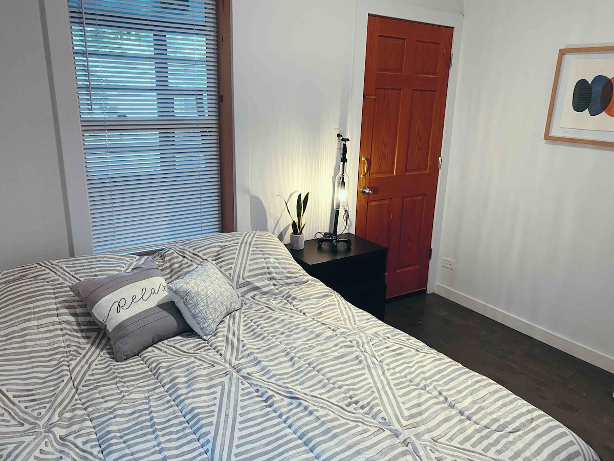 A well-lit bedroom features a spacious bed with a geometric patterned blanket. A bedside table holds a lamp and a small potted plant. Natural light enters through a window with horizontal blinds. A wooden door with a vibrant finish is visible in the background.
