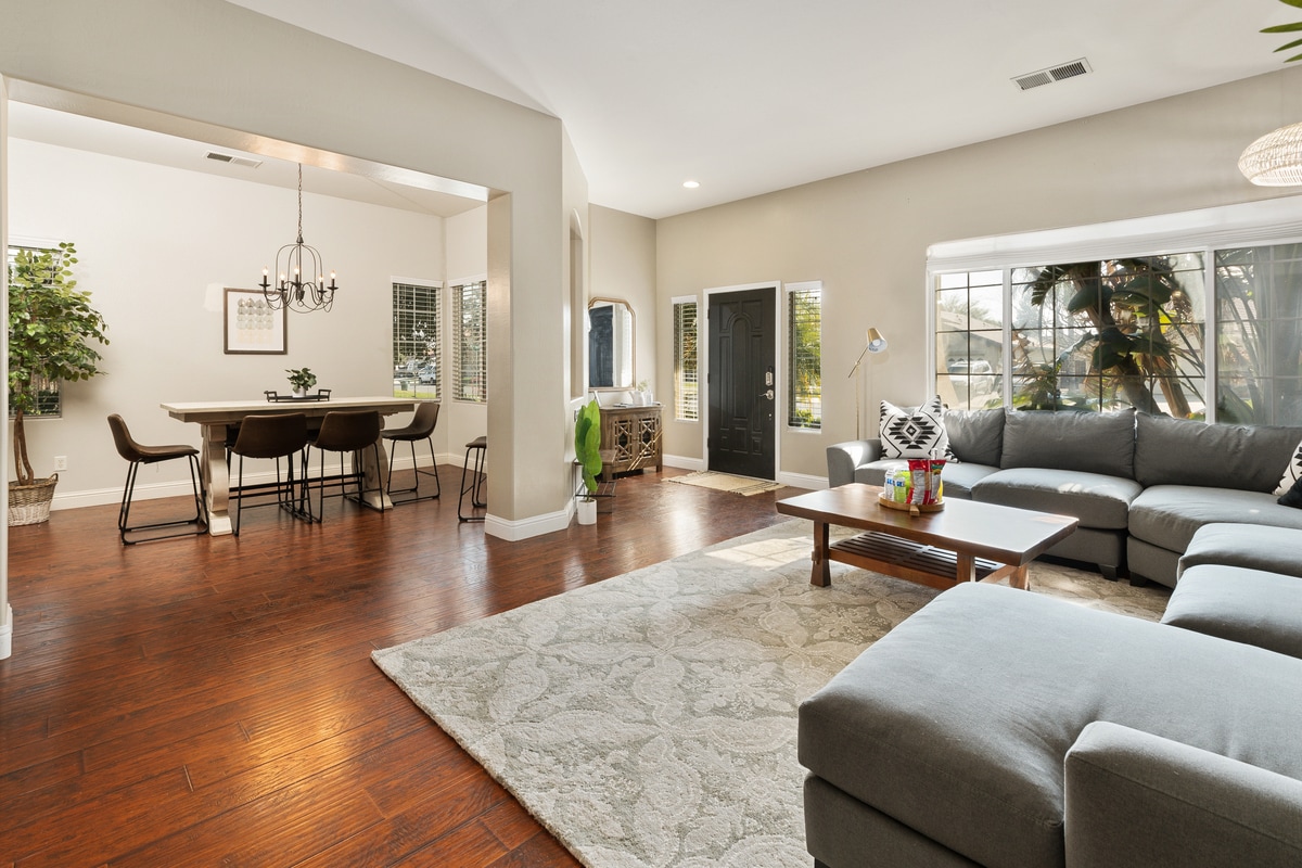 A spacious living area is highlighted by a large sectional sofa and a coffee table, set on a soft area rug. Large windows provide ample natural light, while a dining area with a long table and chairs is visible in the background.