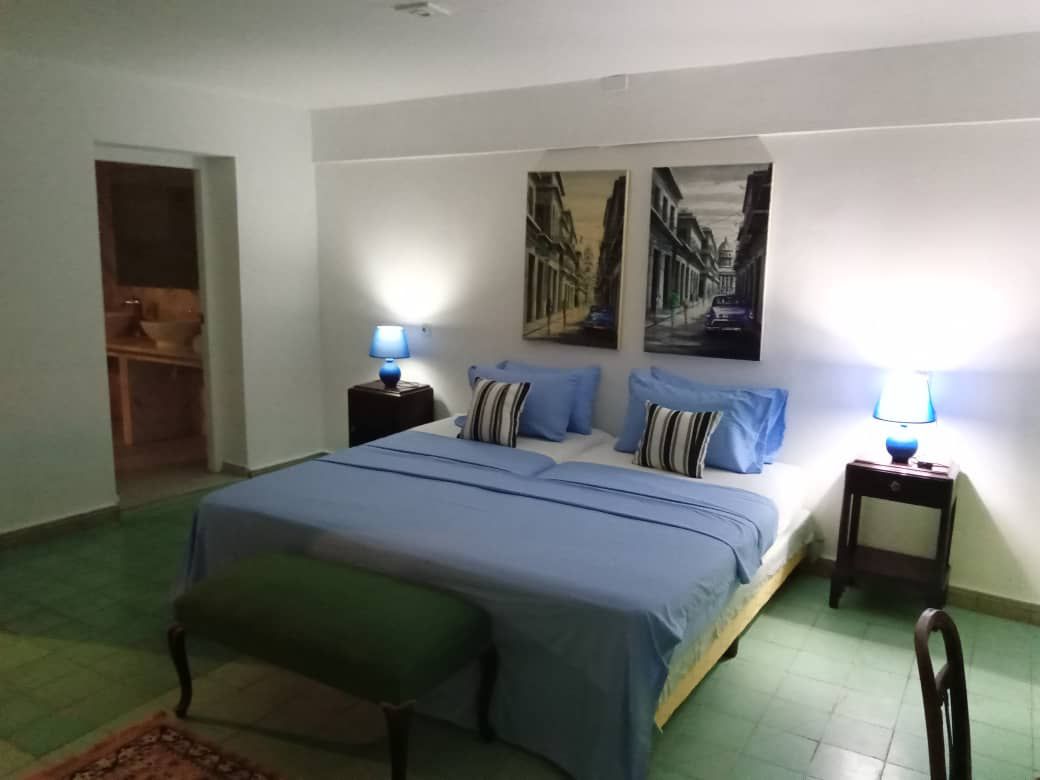 A spacious bedroom is highlighted by a large bed dressed in light blue linens. Two blue lamps illuminate the room, complementing the artwork depicting urban scenes. A green bench sits at the foot of the bed, while the tiled floor offers a cool, calming atmosphere.