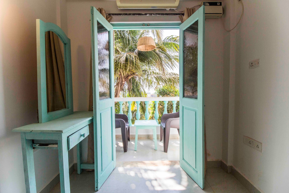 A light-filled room features a pair of open turquoise doors leading to a balcony with a view of palm trees. A simple, matching desk with a mirror is positioned beside the doors, while two comfortable chairs are visible on the balcony.