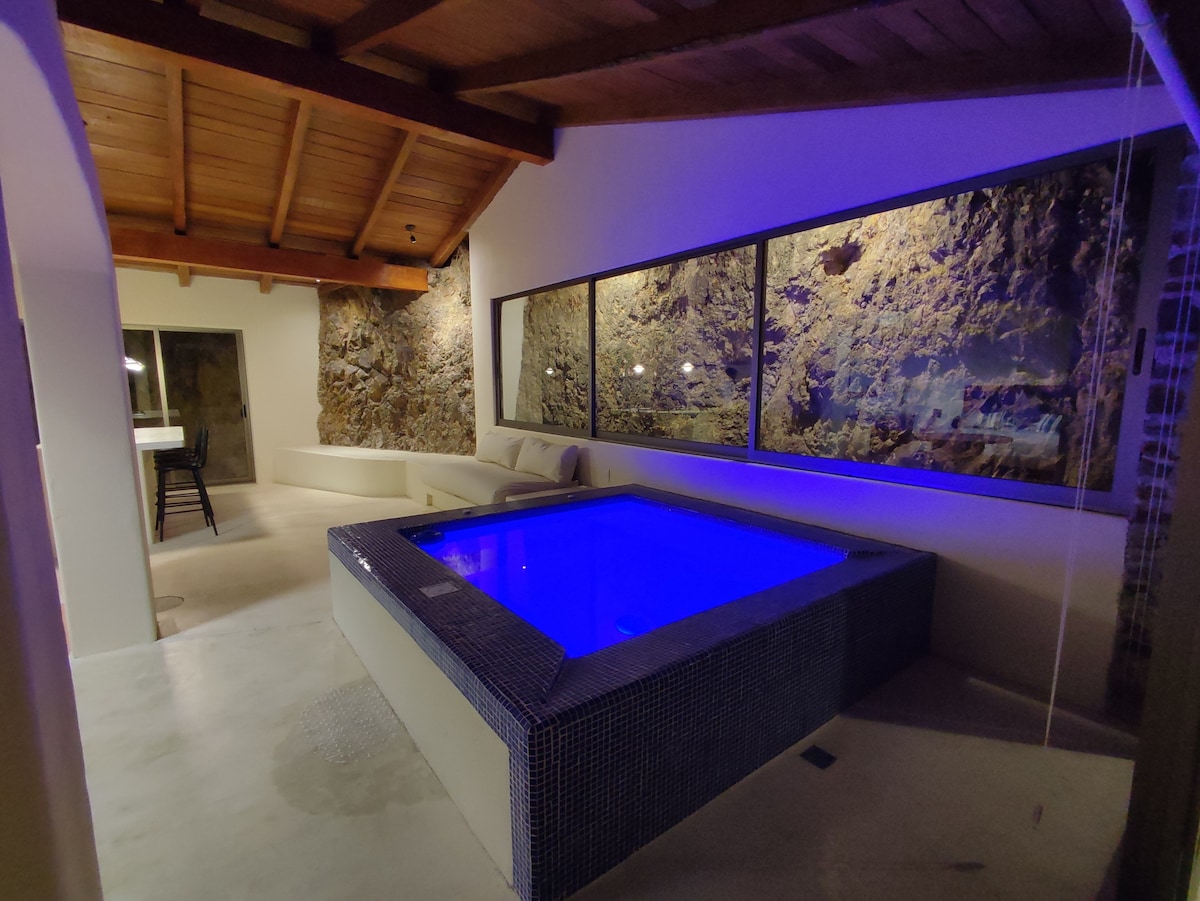 A modern indoor pool is illuminated with soft blue lighting, surrounded by natural stone walls. A seating area with gray cushions is positioned nearby, and large windows provide a view of the outside. The ceiling features wooden beams, enhancing the spacious feel.