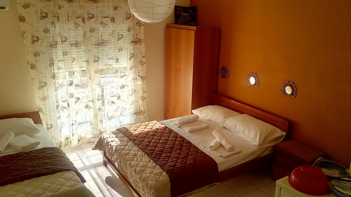 A spacious room is present with two beds, one featuring a quilted coverlet. Natural light enters through curtained windows, which have decorative details. Wooden furnishings include a wardrobe and bedside table, complemented by round wall decorations, creating a cozy and inviting ambiance.
