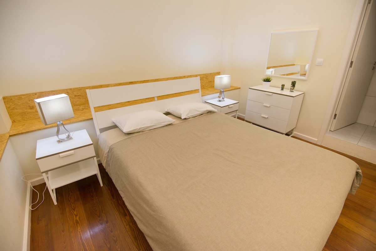 A bright bedroom features a spacious bed covered with a neutral-colored blanket. Two bedside tables with lamps are positioned on either side. A mirror is mounted on the wall, and a dresser is situated nearby, enhancing the room's organized appearance.