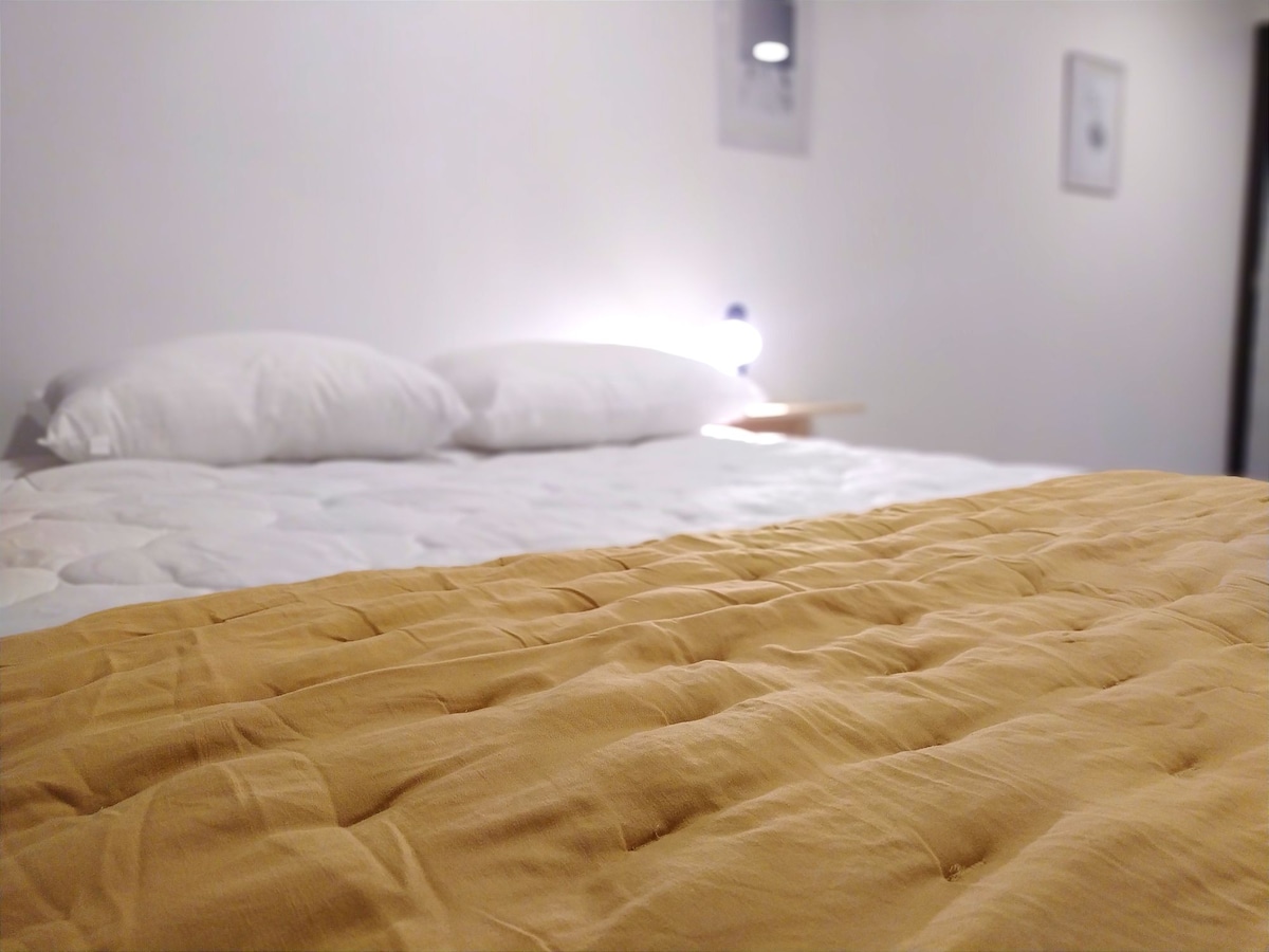 A spacious bed is showcased with a mustard-colored blanket draped over it. Two white pillows rest against a softly lit headboard, creating a calming sleep environment. The background features subtle wall decor, enhancing the simplicity of the space.
