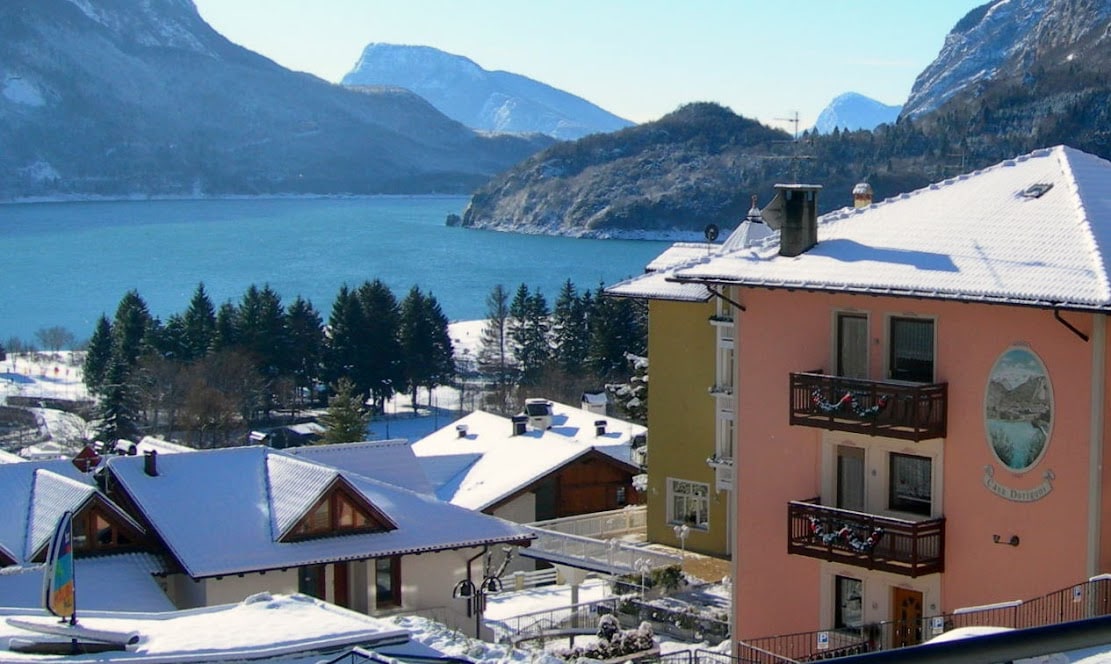 A scenic view captures snow-covered rooftops and the serene landscape of a lake surrounded by mountains. The clear blue water reflects the snowy peaks, while clusters of evergreen trees line the shore, creating a tranquil winter atmosphere.