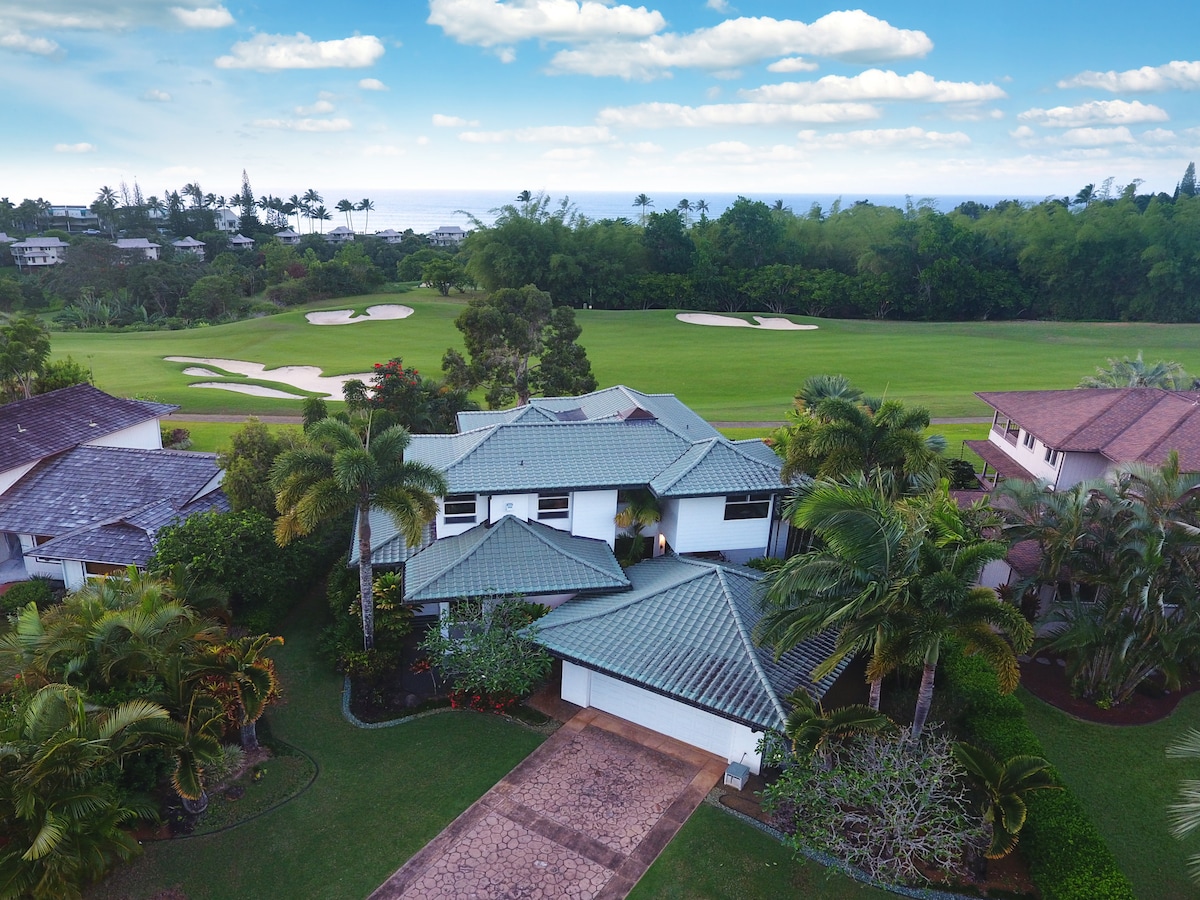 Aerial view of a spacious home surrounded by tropical landscaping. The house features a green-tiled roof and is situated near a golf course, with lush greenery and distant ocean views in the background under a clear sky.