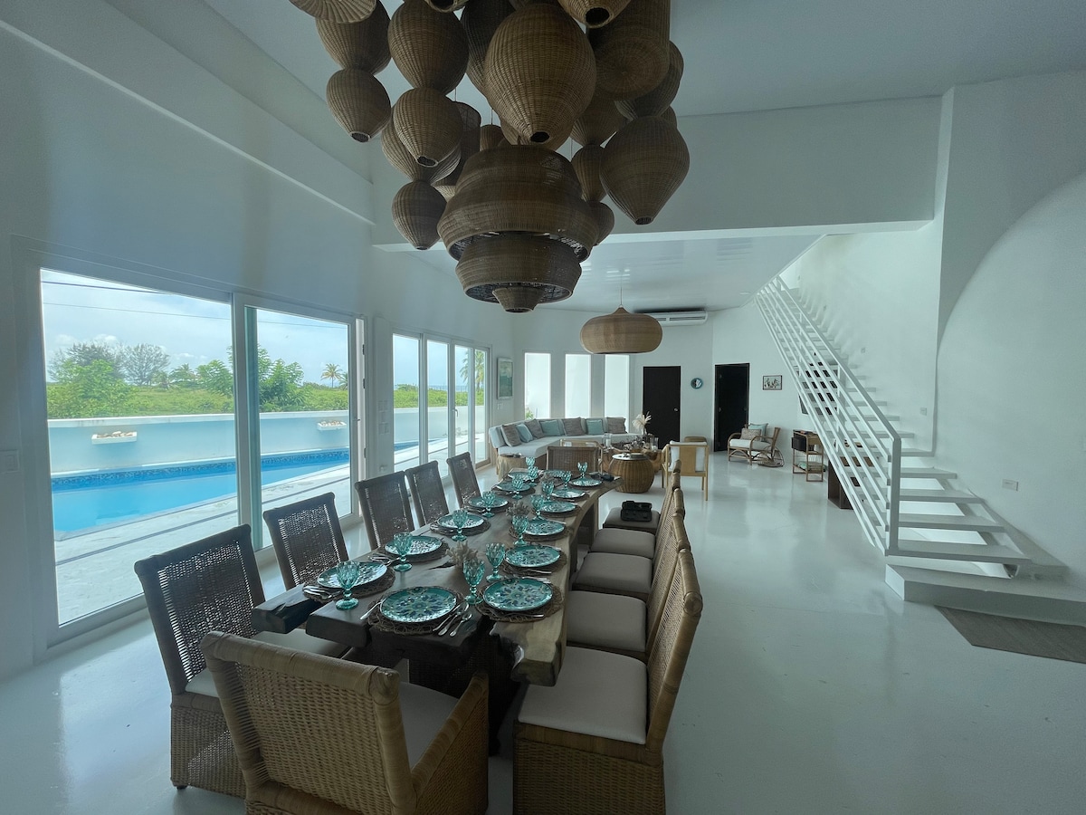 A spacious dining area features a long table set with turquoise plates, surrounded by woven chairs. Large windows provide views of the pool, enhancing the bright and airy ambiance. Light fixtures made from natural materials hang above, contributing to the modern, elegant design.