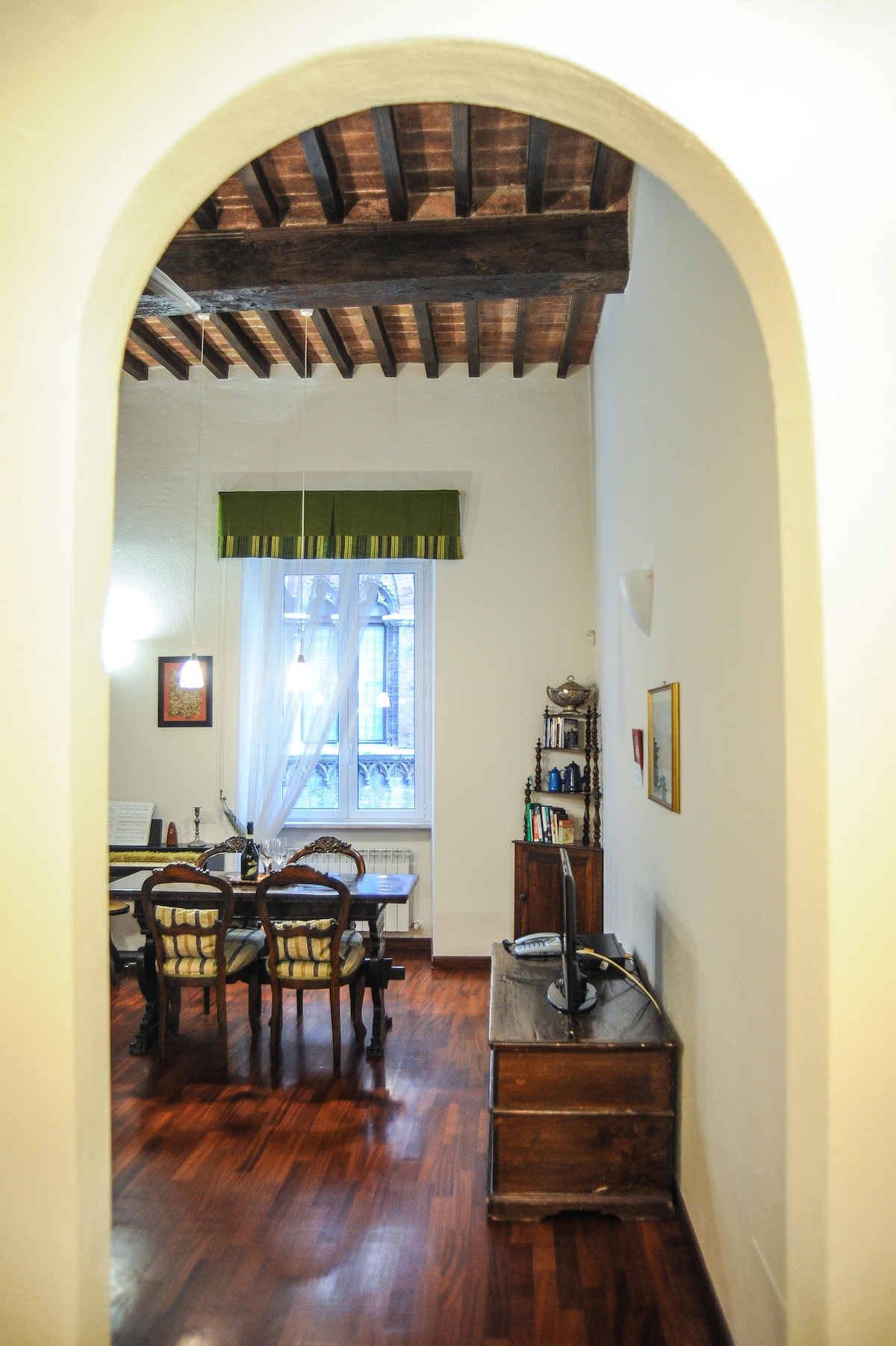 An arched entrance leads into an open-plan living and dining area featuring wooden beams on the ceiling. A wooden table with chairs is visible, along with a cabinet that holds various items. A large window allows natural light to fill the space.