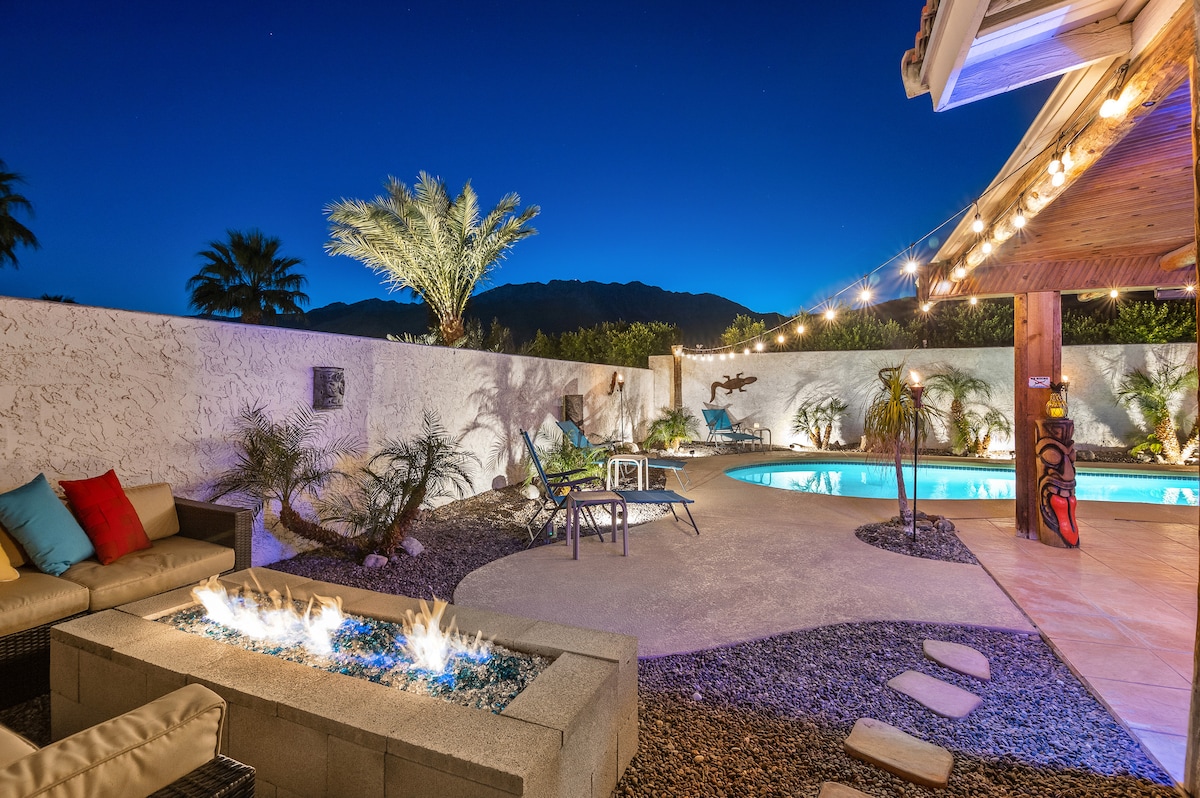 A serene outdoor space is illuminated at night, featuring a crystal-clear pool surrounded by palm trees. A cozy seating area is positioned near a gas firepit. Soft lighting enhances the ambiance, and mountain views are visible in the backdrop.