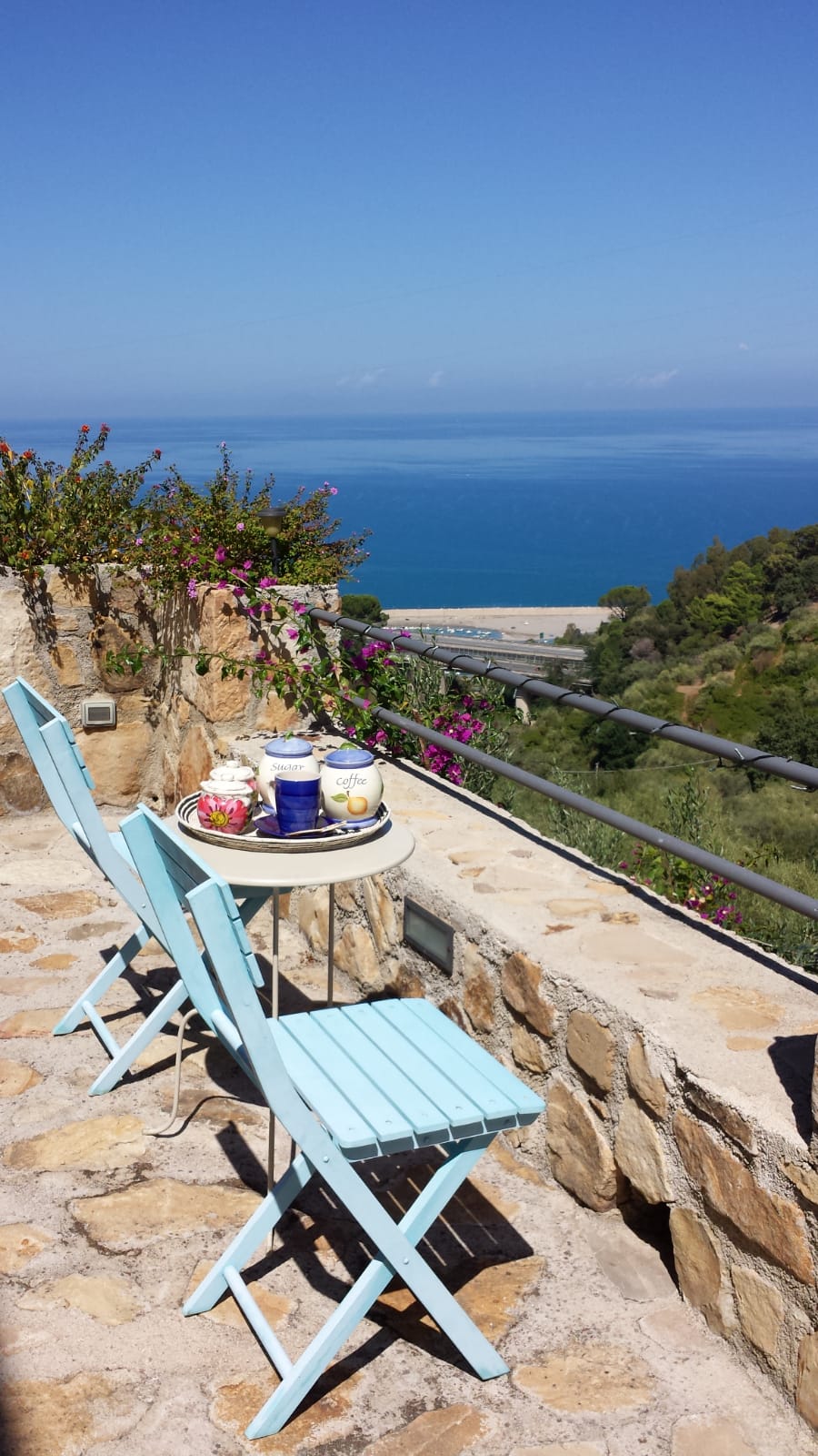 A small outdoor seating area features two light blue chairs and a round table, set against a backdrop of Mediterranean greenery. The table is adorned with tea and small decorative items, while a stunning view of the sea and coastline is visible in the distance.