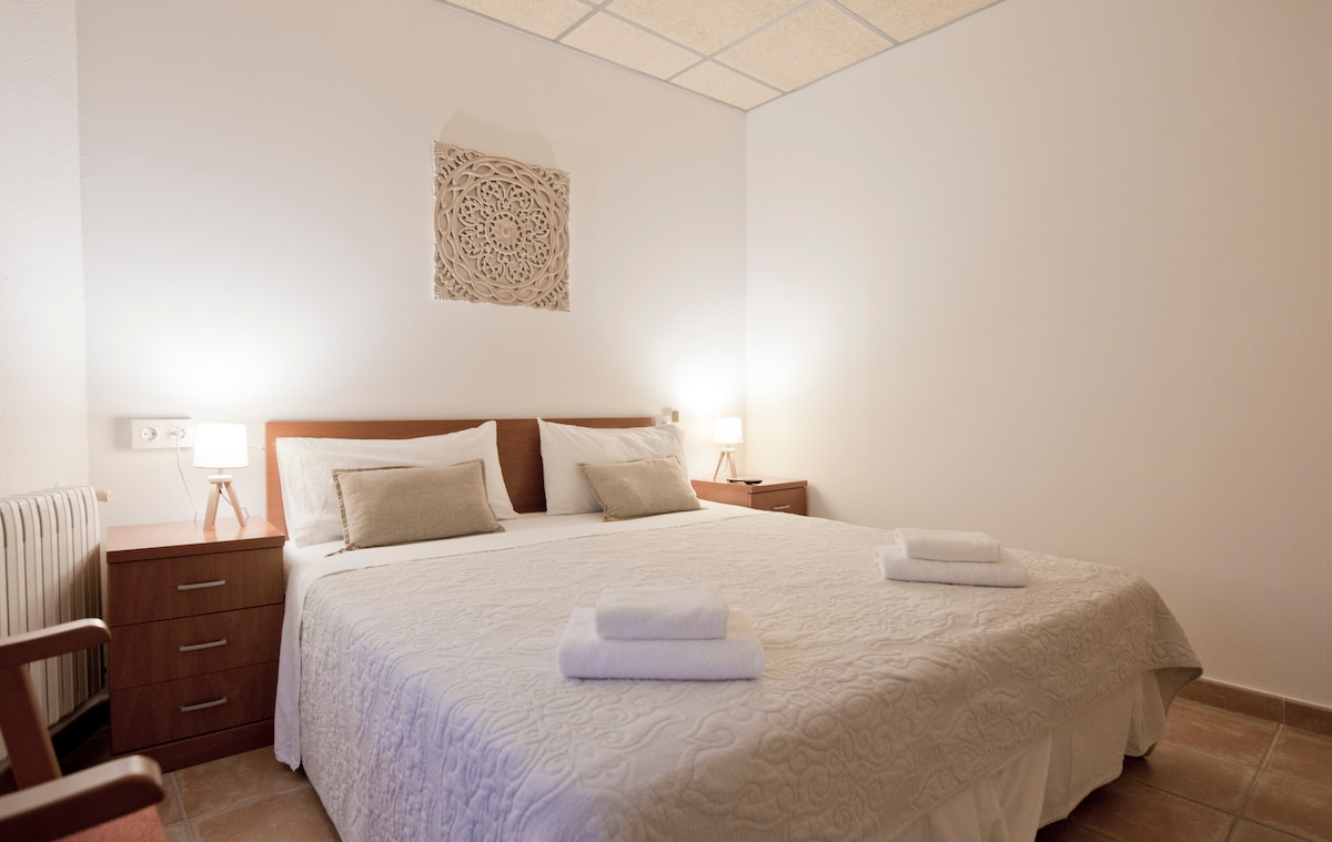 A bright room is showcased with a spacious bed covered in a light blanket. Two neatly arranged towels are placed on the bed. Soft bedside lighting is provided by two lamps on wooden nightstands. A decorative wall piece adds a touch of design to the room.