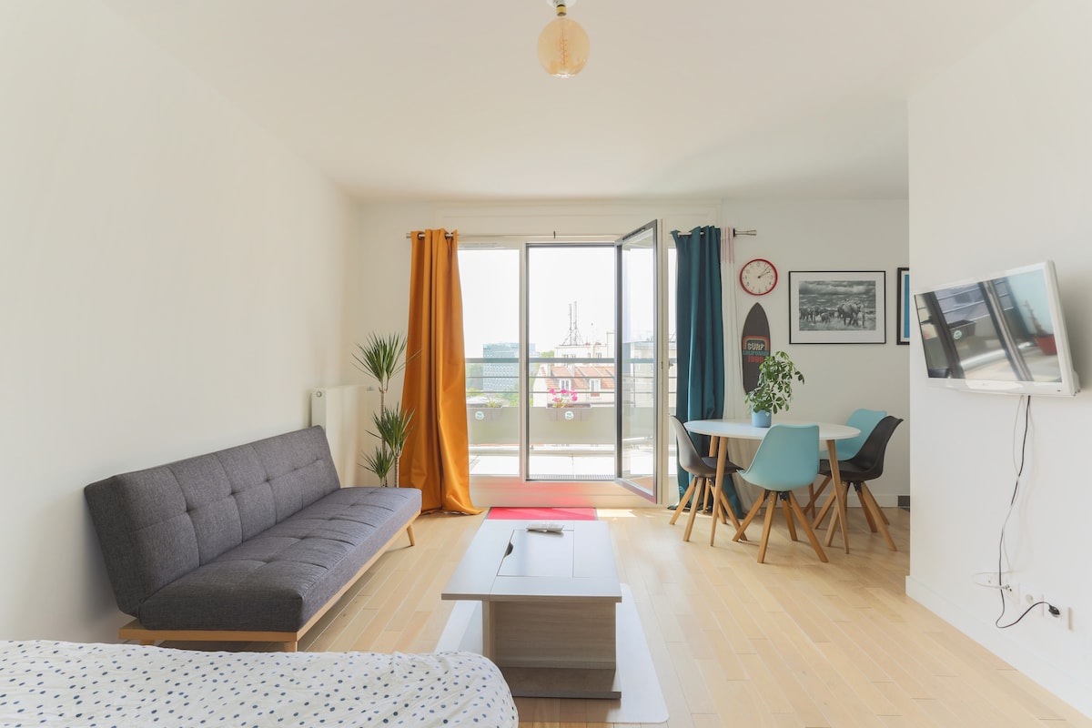 A modern living space features a gray sofa bed and a wooden coffee table, creating a functional area. A dining table with colorful chairs sits nearby, enhancing the room's design. Natural light floods in through a large sliding door that opens to a terrace.