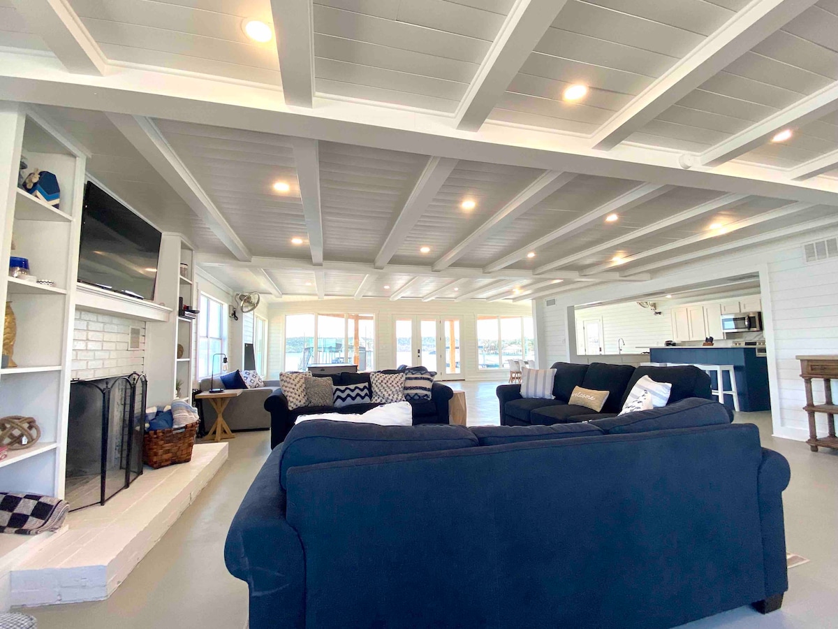 An open living space features a spacious layout with exposed beam ceilings and strategically placed lighting. Comfortable seating options surround a secluded fireplace, while large windows provide natural light and unobstructed views of the nearby lakefront.