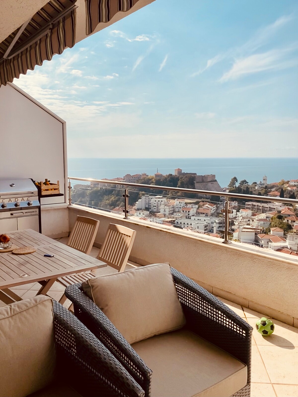 A spacious terrace is visible, featuring a dining table and comfortable lounge seating. A gas grill is positioned nearby. The backdrop showcases a panoramic view of the sea and the fortress, complemented by the distant cityscape bathed in natural light.