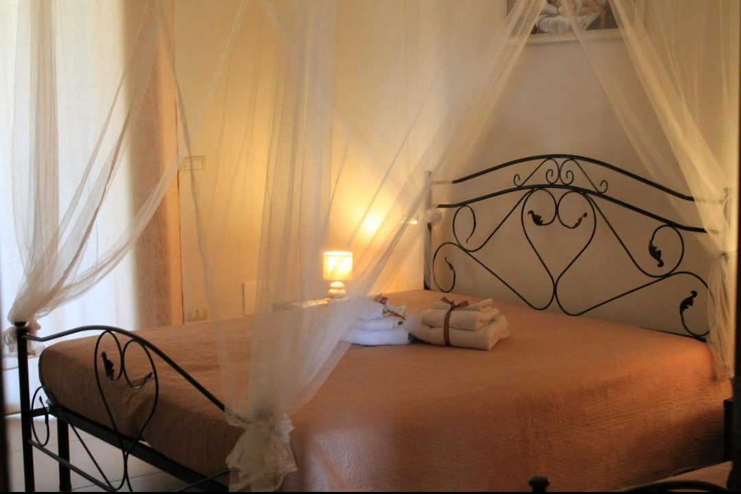 A spacious bed with a wrought-iron frame is dressed in soft linens, surrounded by flowing white drapes. A bedside lamp emits a gentle glow, contributing to a serene ambiance. Freshly laundered towels are neatly arranged at the foot of the bed.
