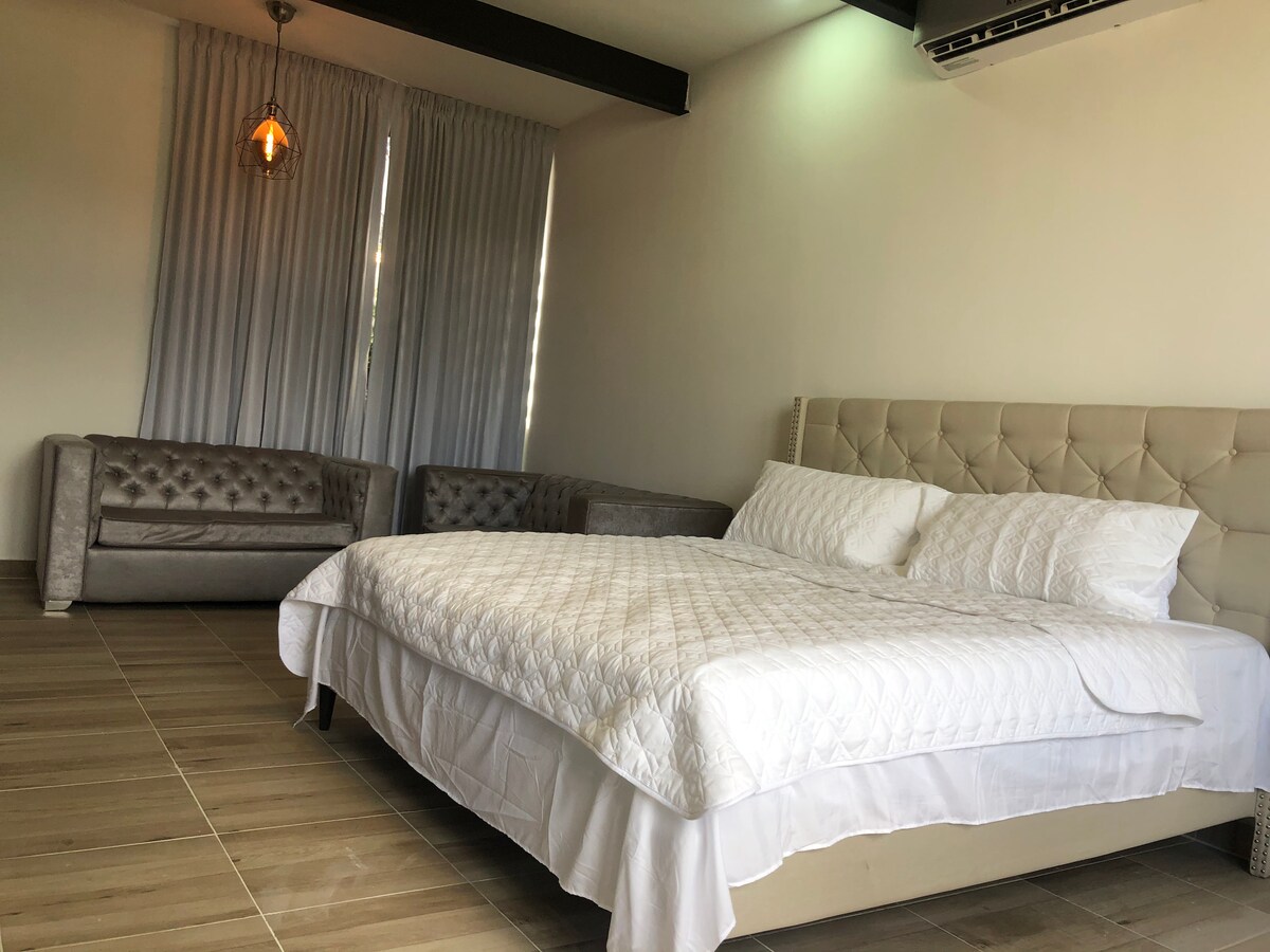 A spacious sleeping area is highlighted by a large, upholstered bed dressed in white linens. Two plush sofas are placed against the wall, framed by sheer curtains that filter natural light, creating a calm environment.