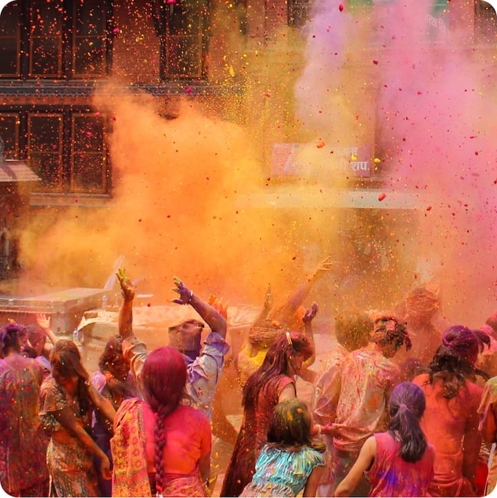 India’s Holi festival. A large group of people, mid-celebration, are seen from behind in the middle of a colorful cloud of pinks, oranges, and purples.