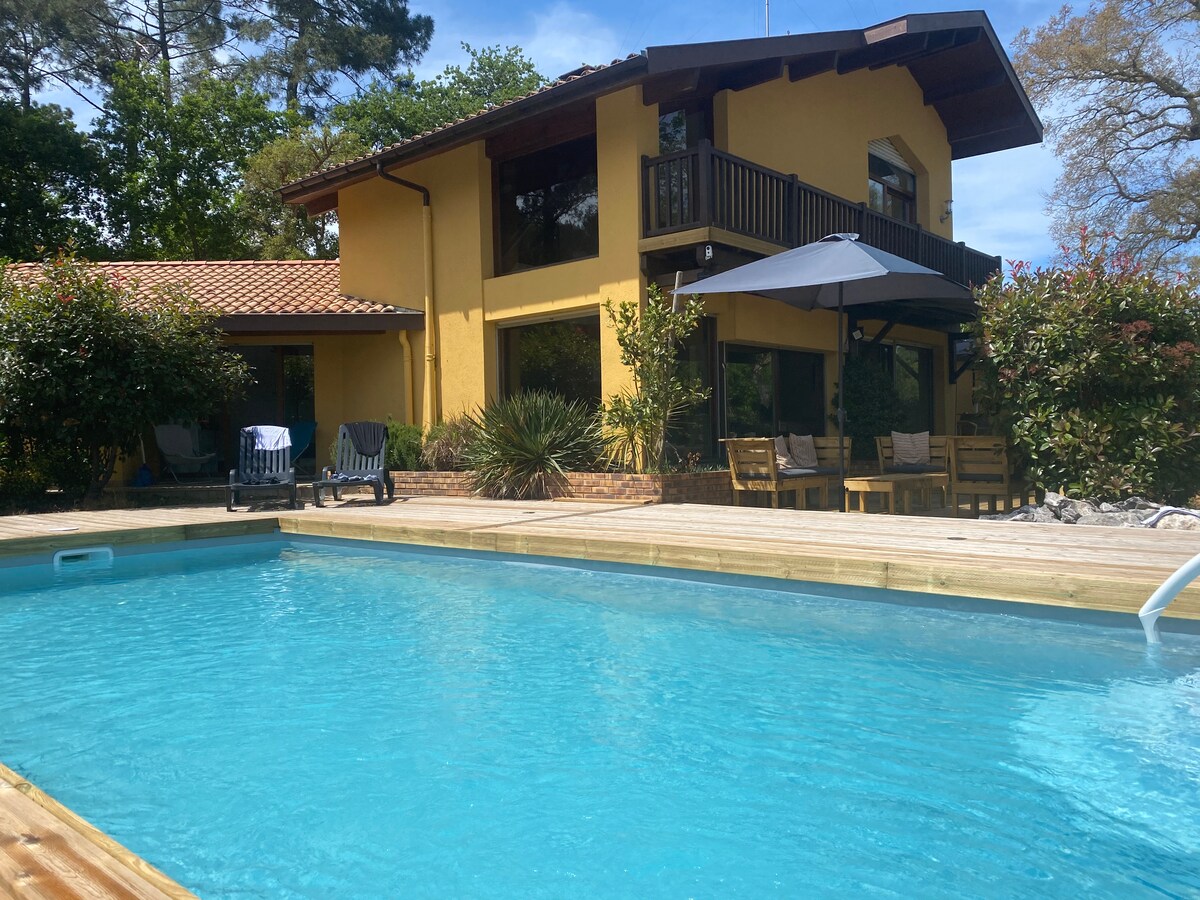 A spacious villa with a vibrant yellow exterior is shown beside a clear swimming pool. Multiple lounge chairs are arranged on the deck, with a large umbrella providing shade. Lush greenery surrounds the area, enhancing the outdoor space for relaxation.