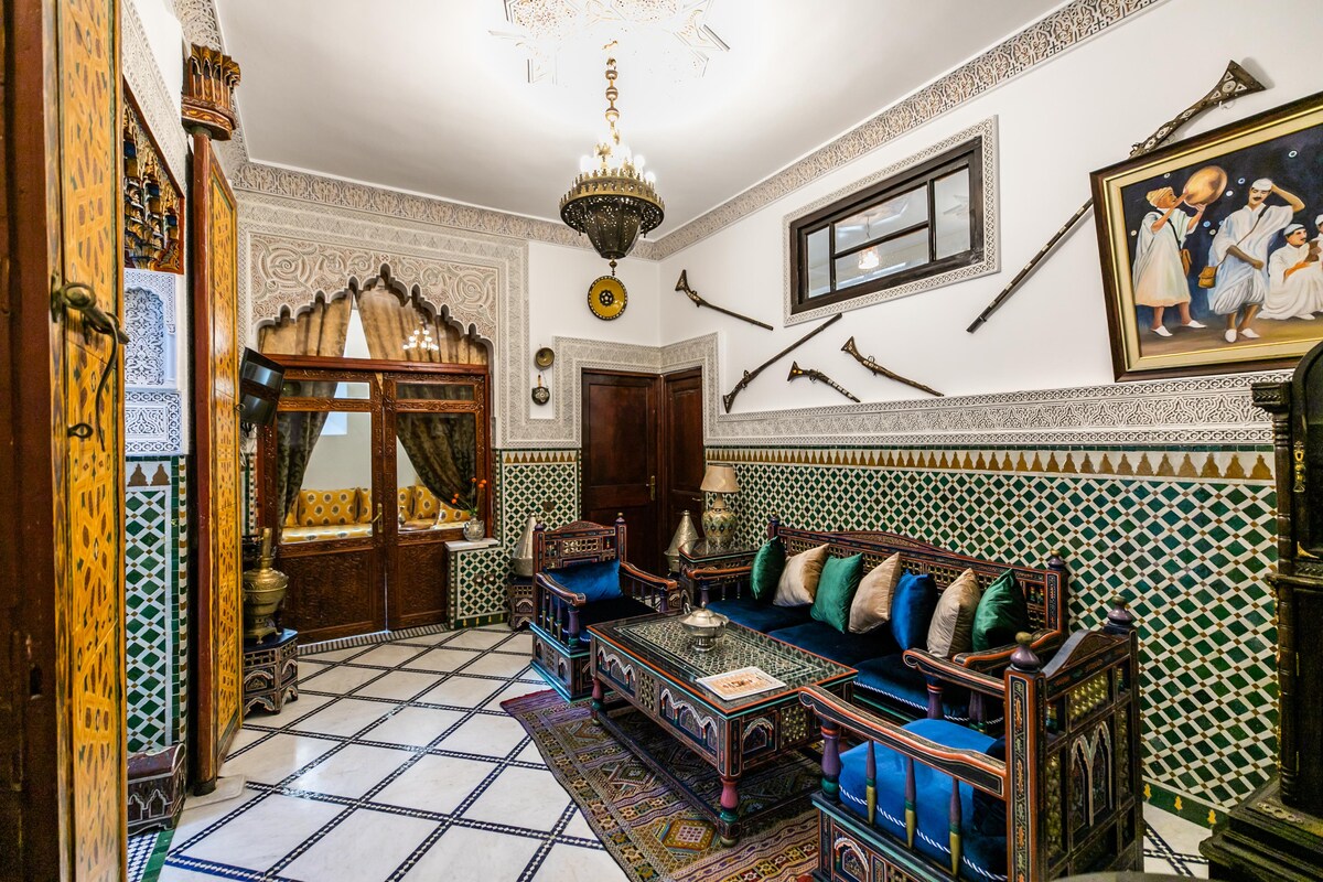 A traditional Moroccan living area is presented, featuring intricately patterned tiles and decorative woodwork. Plush seating in deep blue is arranged around a low table. Ornate chandeliers and framed artwork reflect the local culture, while decorative weapons are displayed on the walls.