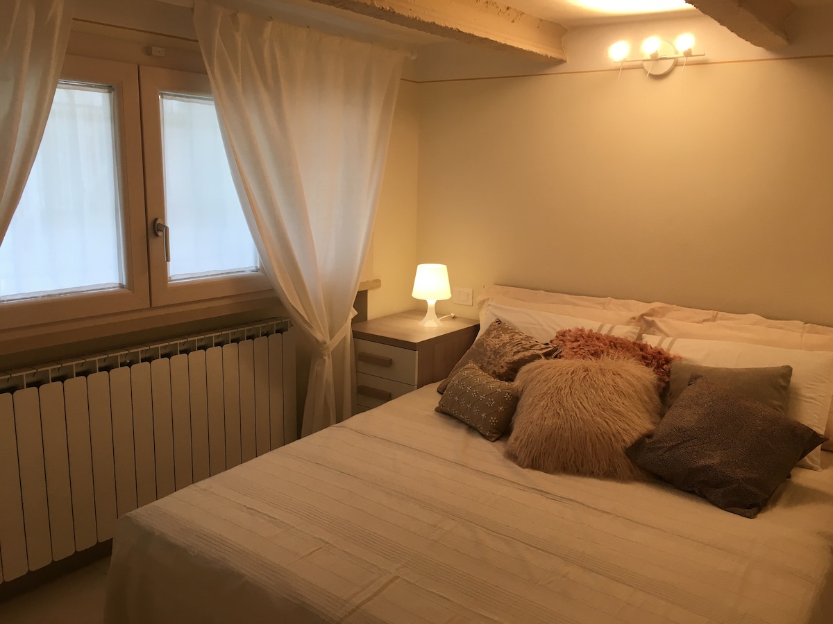 A cozy sleeping area features a spacious bed with soft pillows and a textured throw. Two windows with light curtains allow natural light to enter, while a bedside lamp provides additional illumination. A radiator is located below the windows, enhancing comfort.