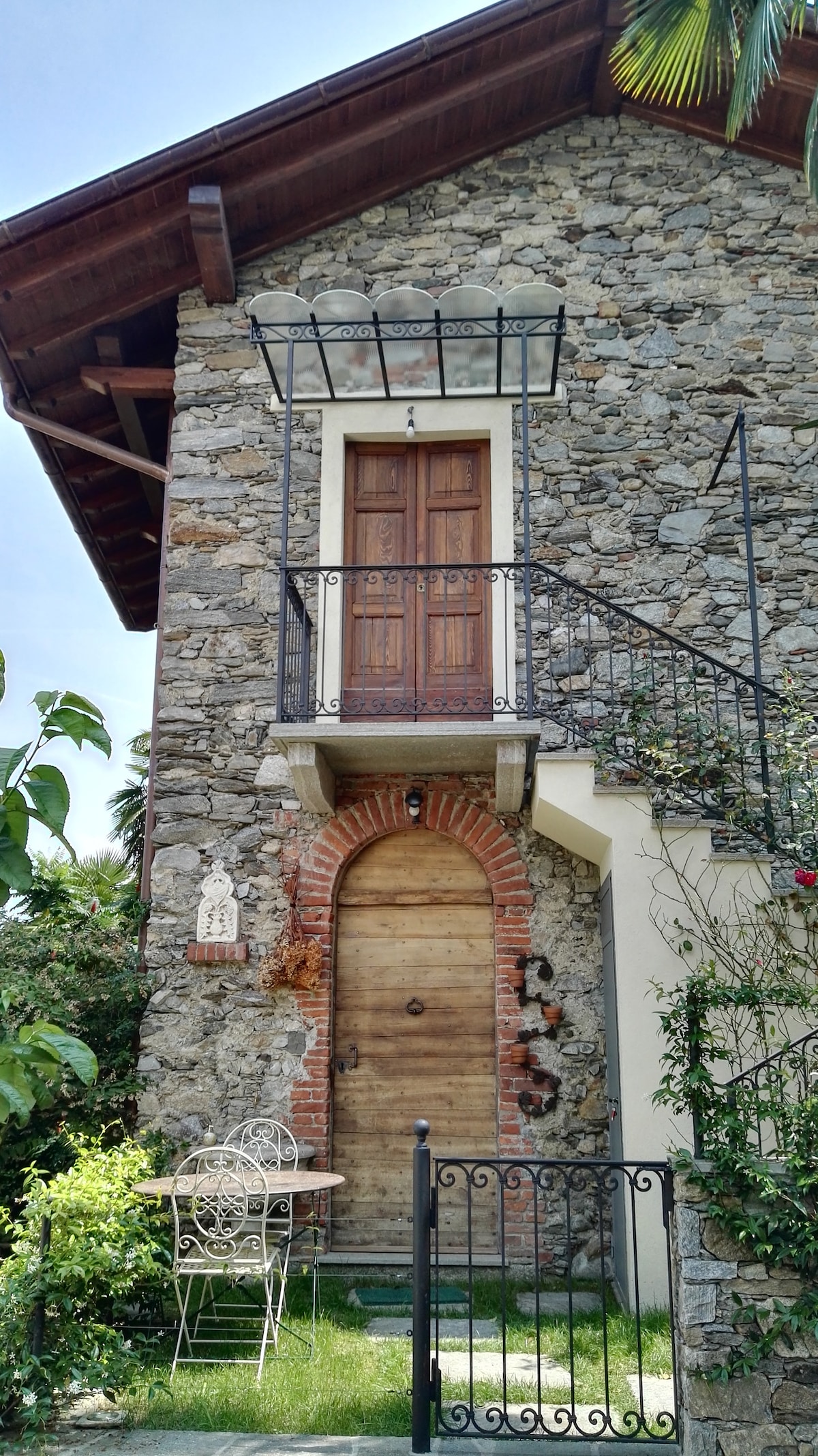The exterior features a charming stone façade with an intricate iron balcony. A wooden door with distinctive carvings is centered, flanked by a small decorative entrance. A quaint seating area with a table and chairs is nestled beside vibrant greenery, inviting relaxation in the serene surroundings.