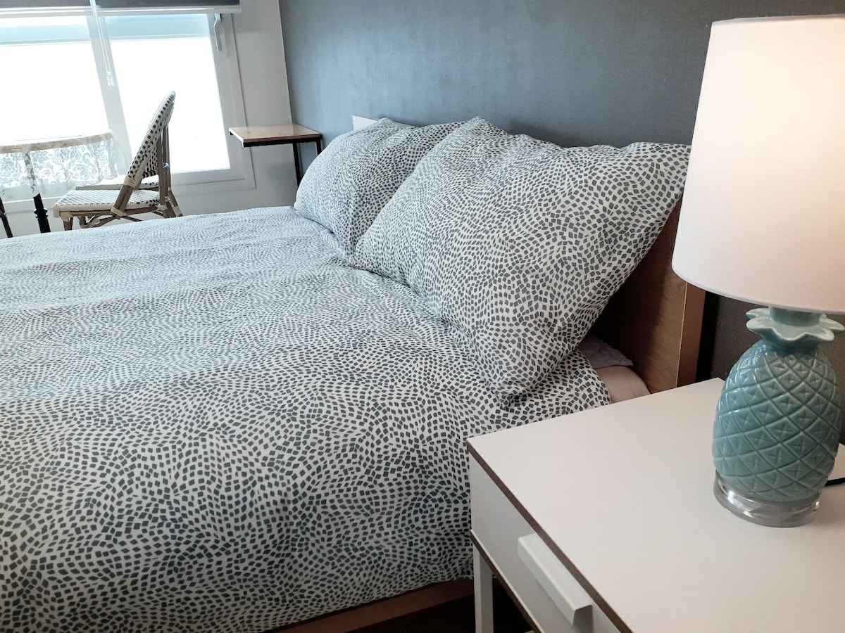 A spacious bed is adorned with a patterned duvet and matching pillows. A bedside table with a lamp is positioned next to the bed, while a chair and a small table are visible in the background. Natural light filters through the window.