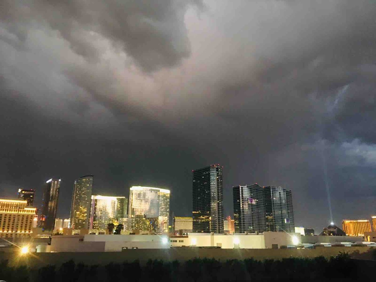 A city skyline is visible against a dramatic sky filled with dark clouds. Bright lights from buildings illuminate the scene, showcasing a vibrant contrast with the stormy backdrop. The urban landscape features a mix of modern architecture and illuminated structures, enhancing the evening view.
