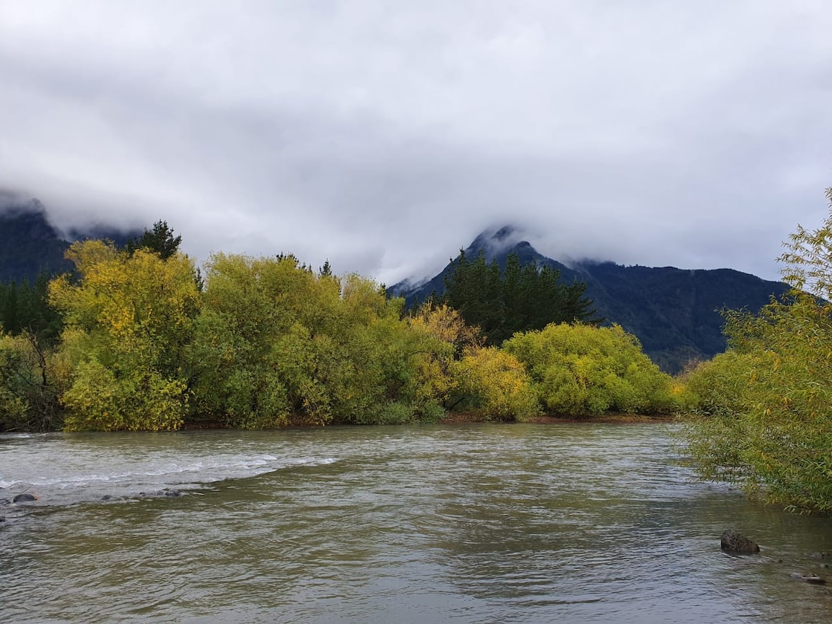 A serene river flows through a landscape adorned with lush greenery and trees displaying autumn colors. Dark clouds hover over the mountains in the background, creating a peaceful yet dramatic atmosphere.