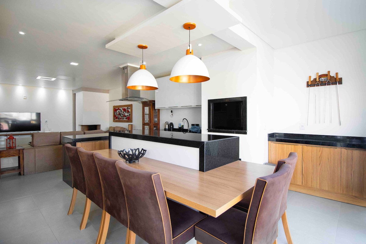 A modern kitchen and dining area is presented, featuring a spacious wooden dining table surrounded by brown upholstered chairs. Two pendant lights illuminate the area. The kitchen includes sleek black countertops and stainless steel appliances, with an open layout connecting to a living space.
