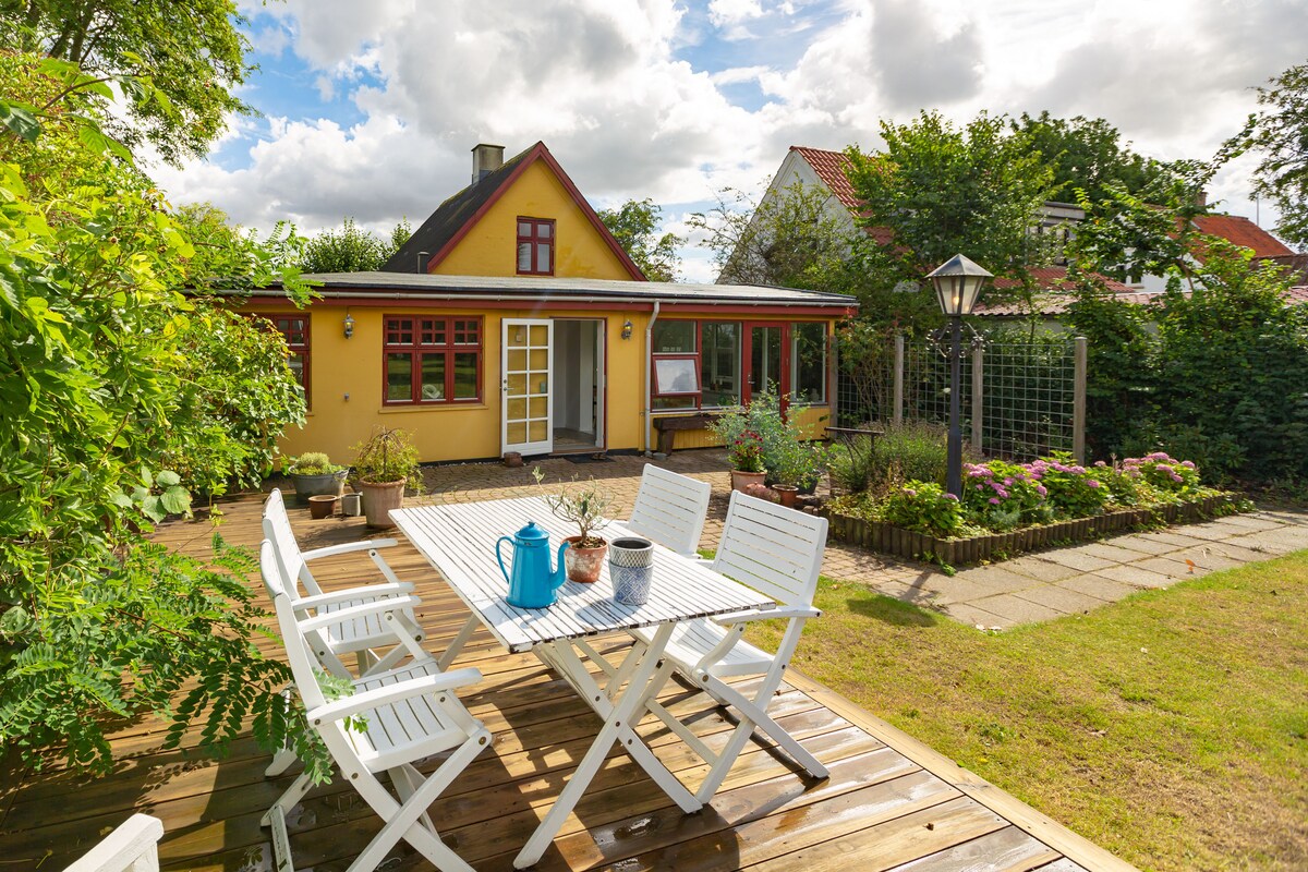 An outdoor deck area is furnished with a white dining table and chairs, surrounded by greenery and colorful flowers. Sunlight highlights the cozy yellow house with large windows and a welcoming entrance. A garden lamp stands nearby, enhancing the inviting outdoor setting.