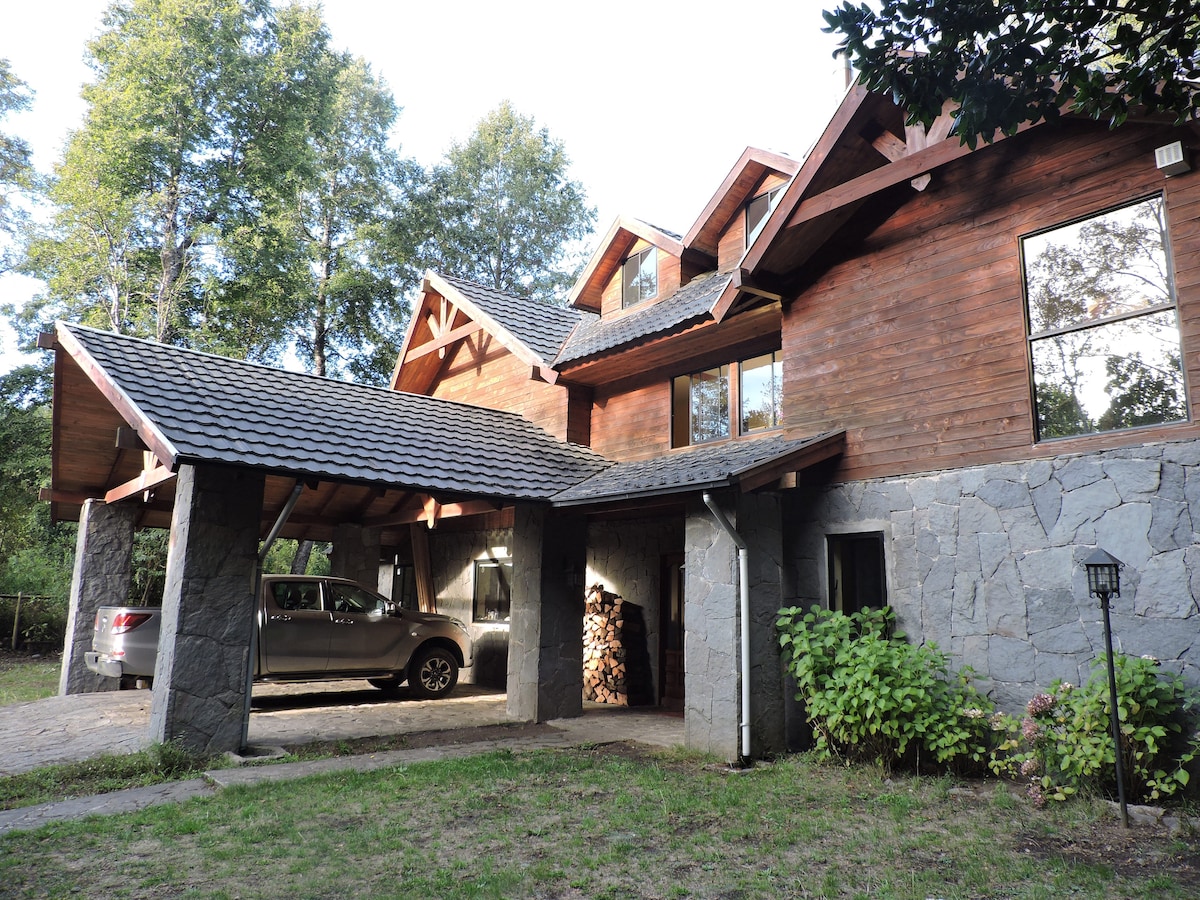A wooden house features a stone foundation and large windows, allowing natural light to illuminate the interior. A covered porch offers a view of the surrounding greenery, with a vehicle parked nearby and firewood neatly stacked. Lush plants add to the serene landscape.