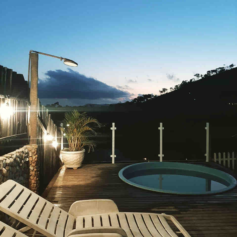 An inviting pool area surrounded by wooden decking is illuminated softly by outdoor lights. Sun loungers provide a comfortable space for relaxation, while a gentle slope of greenery in the background offers a peaceful view against the evening sky.