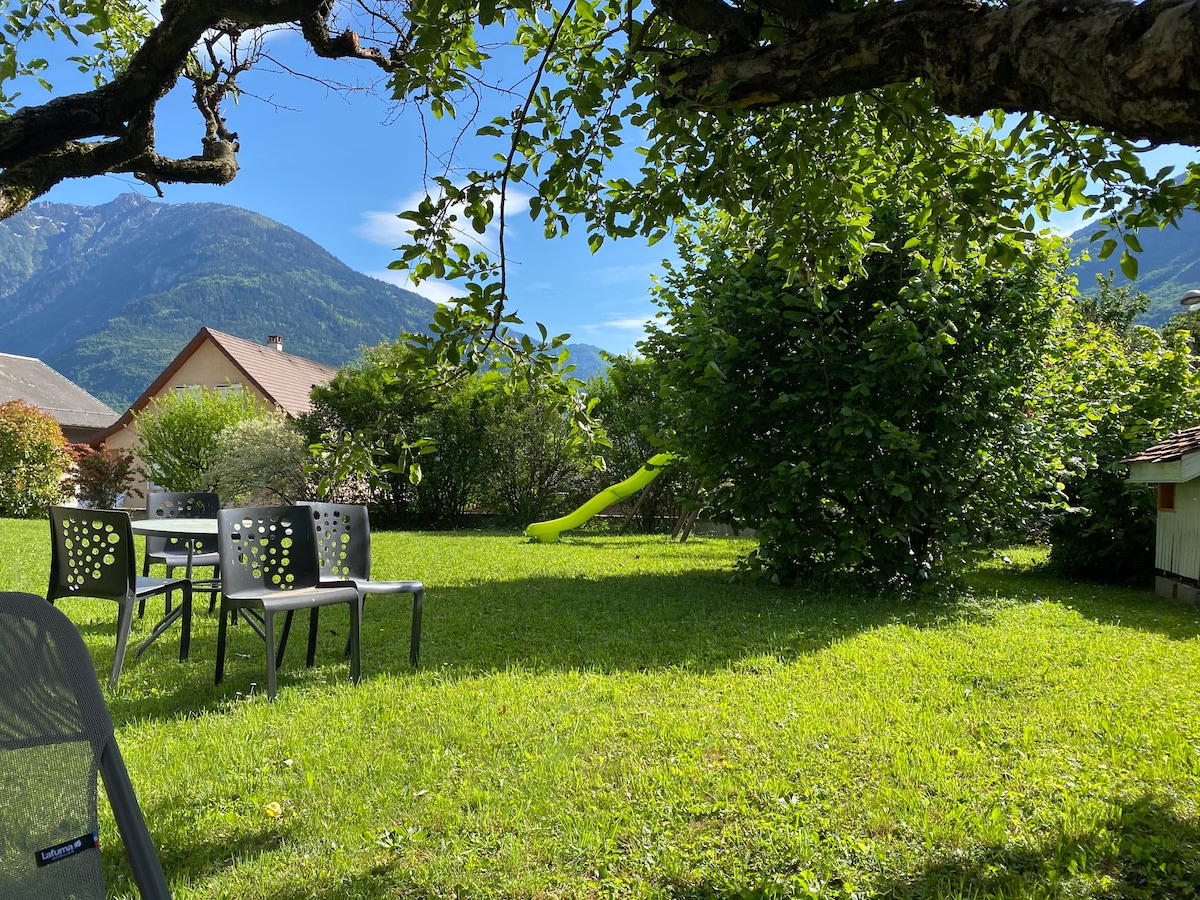 A green garden is featured, with a few gray chairs arranged around a table on the grass. A vibrant green slide is positioned nearby, surrounded by trees and shrubs. Rolling mountains are visible in the background under a clear blue sky.