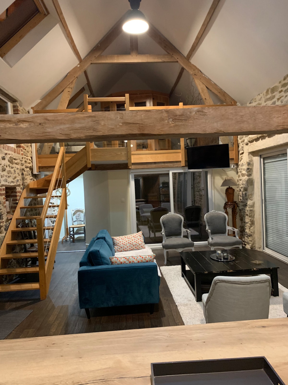 An open living space showcases a high ceiling with exposed wooden beams. A blue sofa is positioned beside a black coffee table, complemented by four armchairs. A staircase leads to a loft area, enhancing the room's spaciousness. Large windows allow natural light to enter.