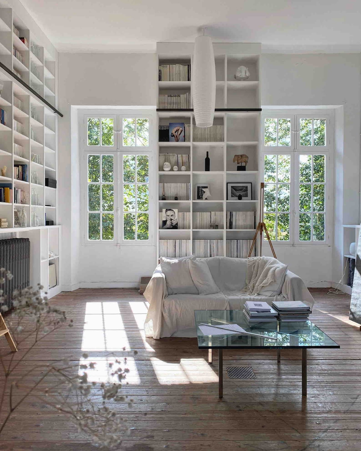 A bright living space features large windows allowing natural light to illuminate a white sofa draped with a light throw. Bookshelves filled with books and decorative objects line the walls, complemented by a glass coffee table adorned with neatly stacked materials.