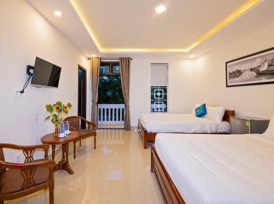 A spacious room is furnished with two beds, a seating area with a wooden table, and a flat-screen TV mounted on the wall. Natural light filters through the windows, which are dressed with light curtains. A refrigerator is discreetly placed in the corner.