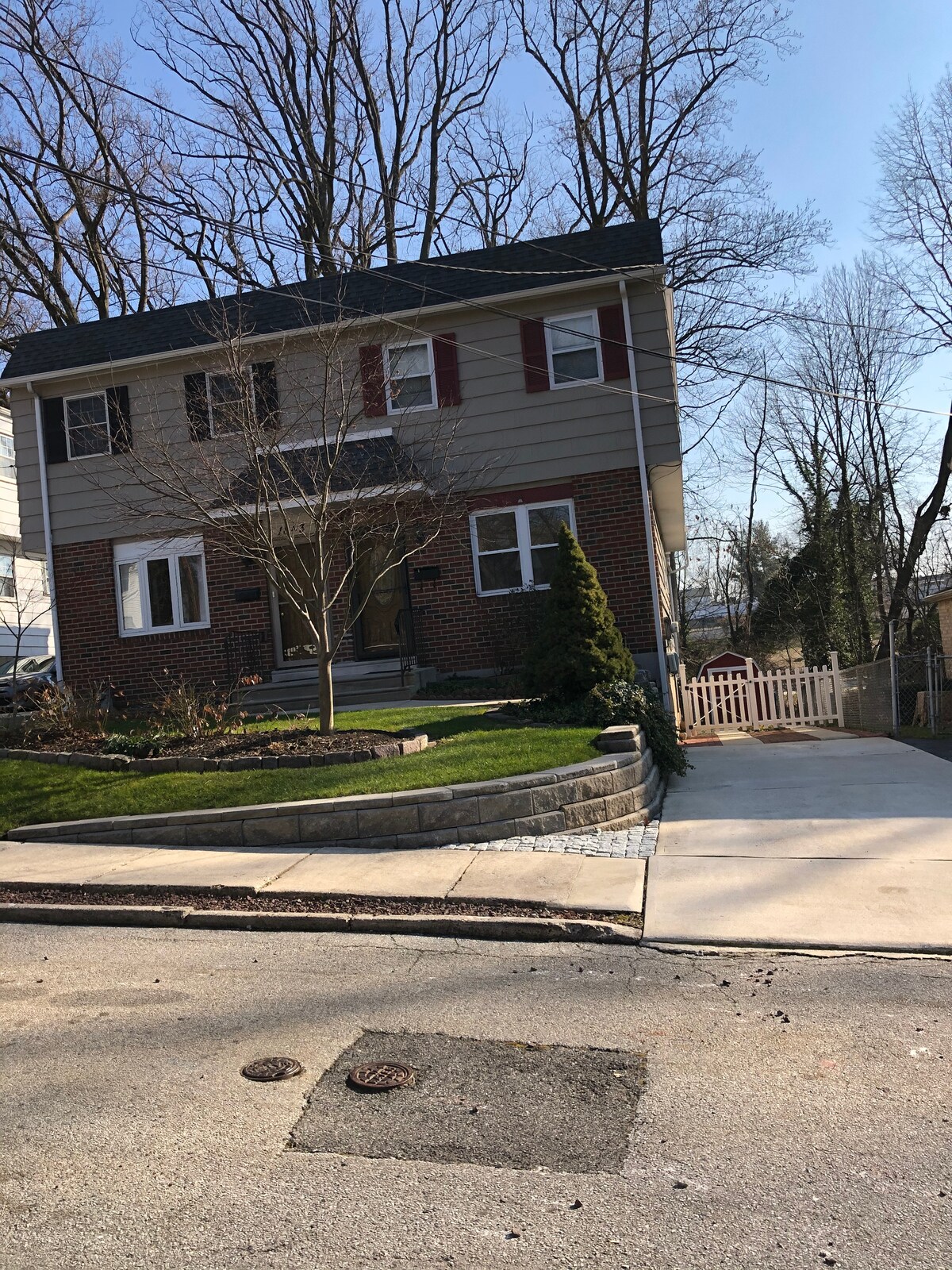 A two-story twin townhouse is depicted, featuring a brick and siding exterior. A landscaped yard includes a small tree and entrance steps leading to the main door. A concrete driveway and sidewalk provide access, with a white picket fence visible in the background.