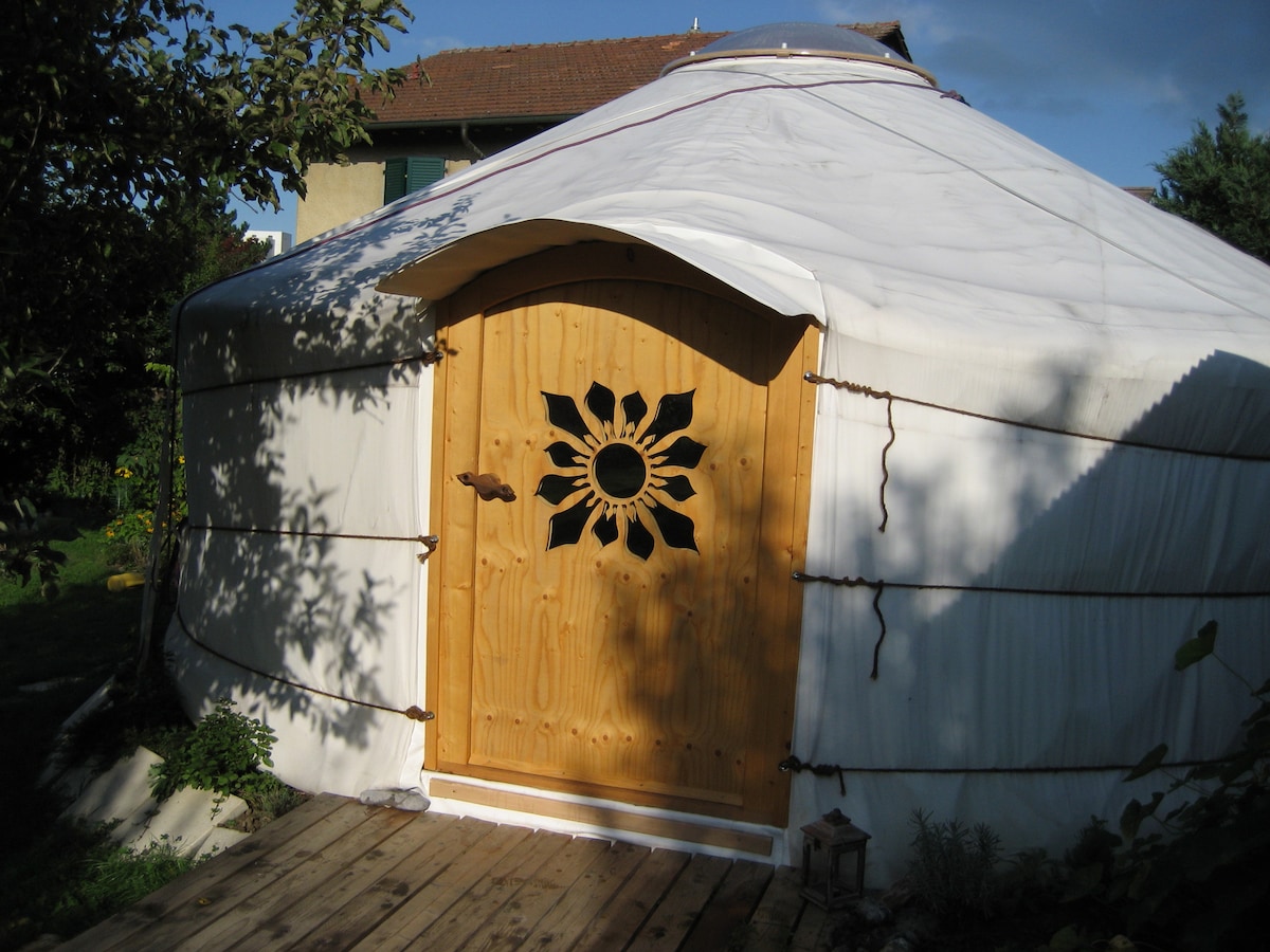 A traditional yurt features a wooden door with intricate floral cut-out designs. The structure is surrounded by greenery, with a wooden pathway leading to the entrance. The exterior is white and is complemented by a peaceful setting that reflects the natural surroundings.