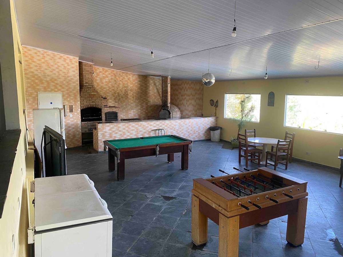 A spacious party room features a billiards table and a foosball table, set on a stone-tiled floor. Large windows provide ample natural light, while a brick fireplace and an outdoor pizza oven are present in the background. Seating is arranged with several wooden chairs around a central table.